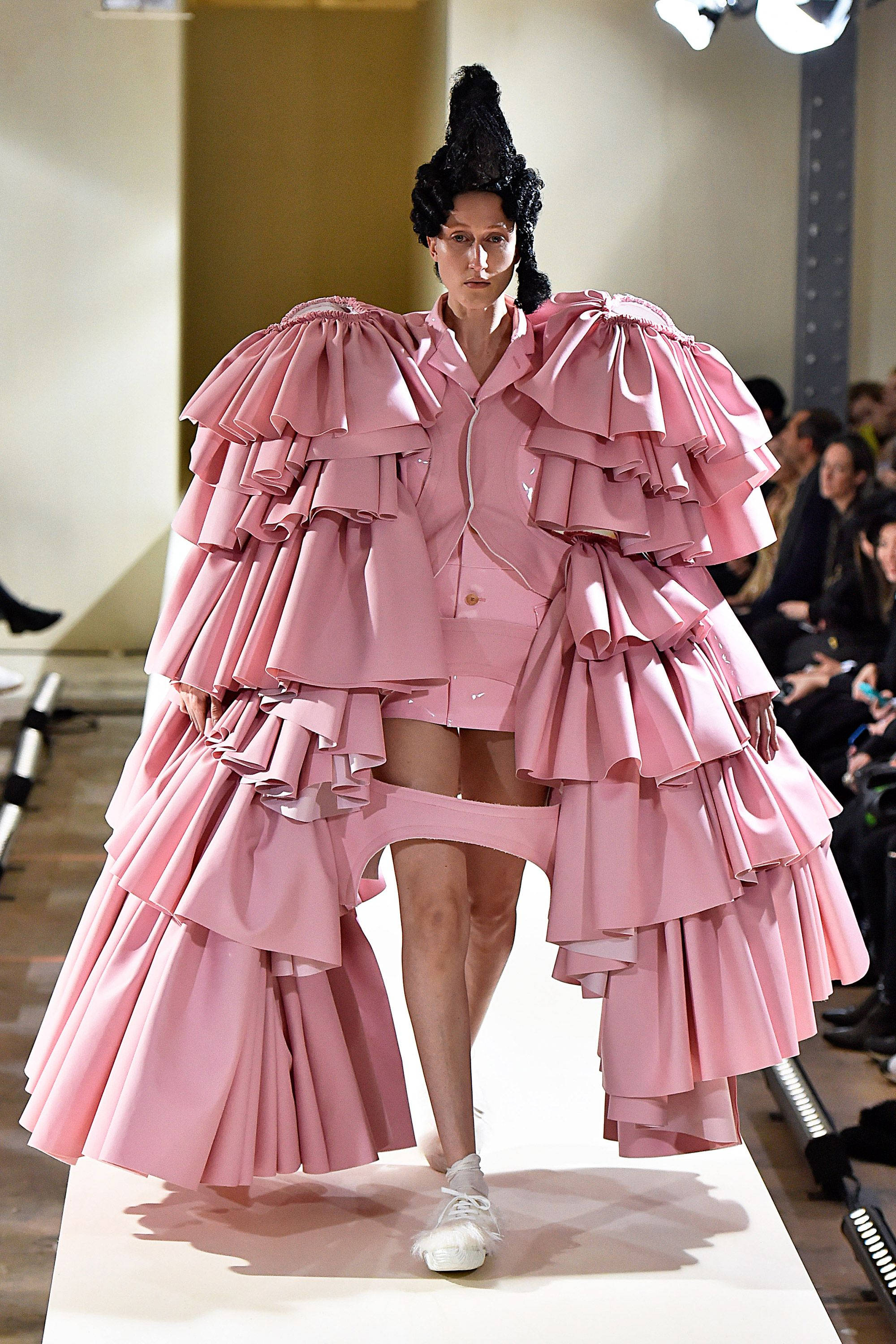 Comme Des Garçons 巨型粉色荷葉邊連衣裙 壁紙 — 一位模特兒穿著前衛的粉紅色荷葉邊服裝，搭配黑色高聳頭飾和白色運動鞋走在天橋上。