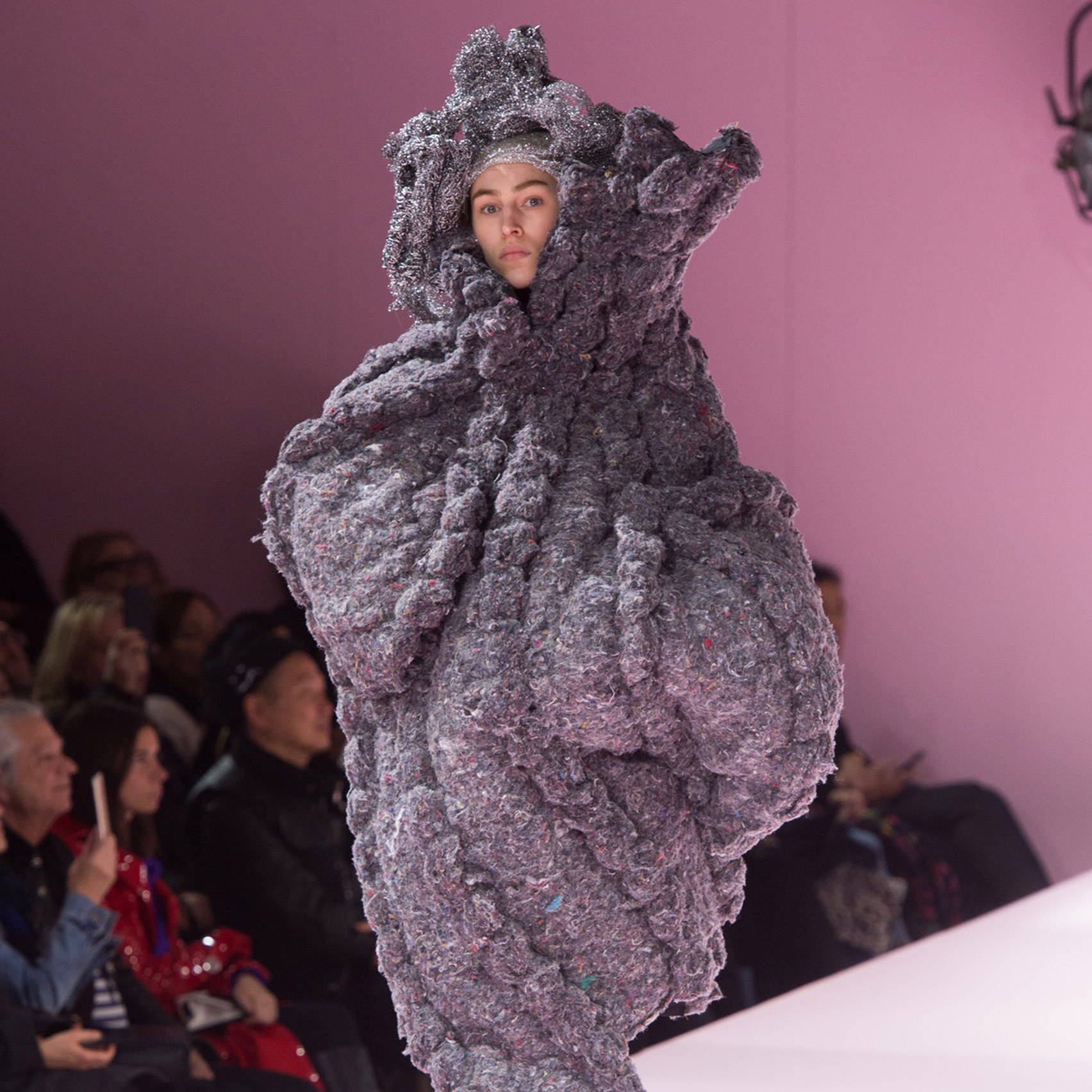 Comme Des Garçons 復用布料廢棄物——創新時尚方法 壁紙 — 一位模特兒身穿帶有雕塑感帽子的前衛紋理灰色服裝，在粉紅色背景的T台上走秀。