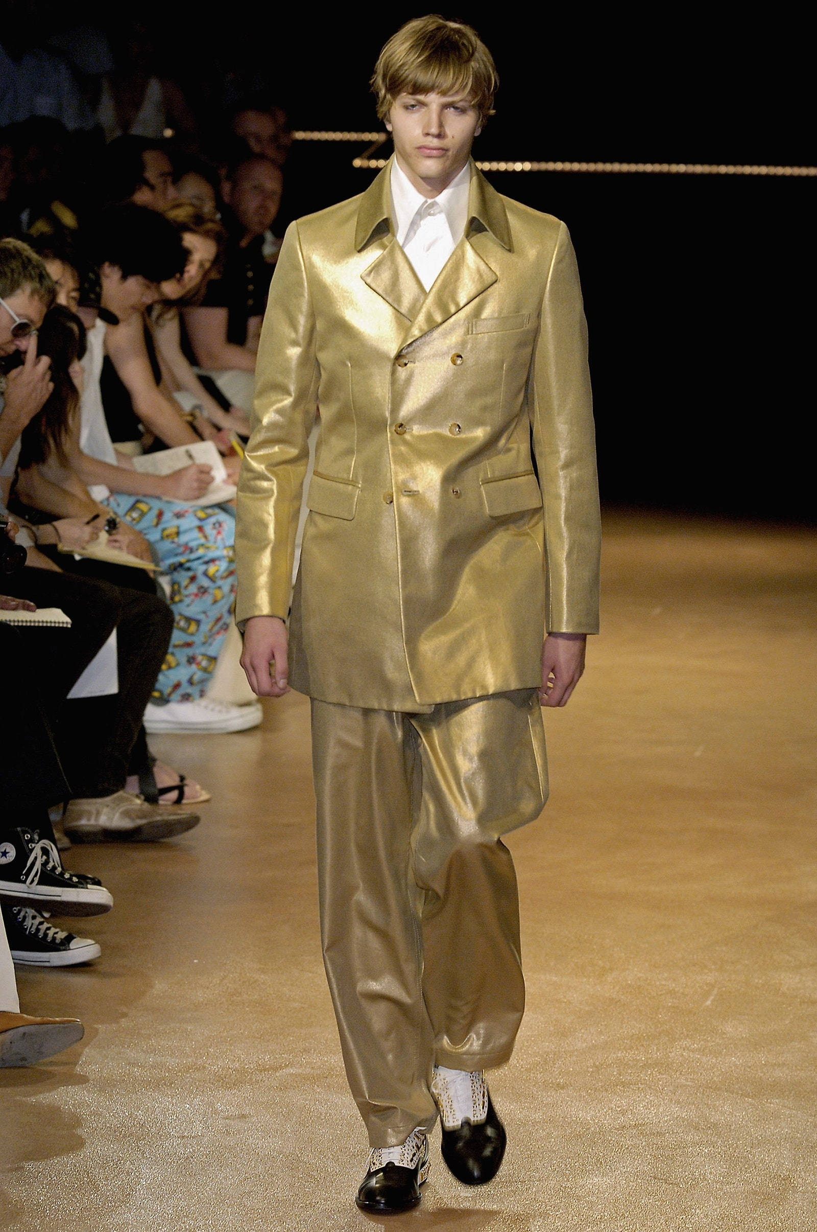 Comme Des Garçons 金色西裝 壁紙 — 一位模特兒穿著閃亮金色雙排扣西裝，搭配寬鬆長褲和黑色鞋子，在時裝展的舞台上走秀。