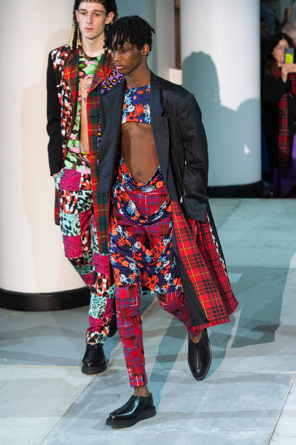 Comme Des Garçons花卉撕裂上衣 壁紙 — 兩位男模特兒自信地走在天橋上，身穿黑色外套下大膽色彩繽紛的拼布服裝，帶有格紋和花卉圖案。