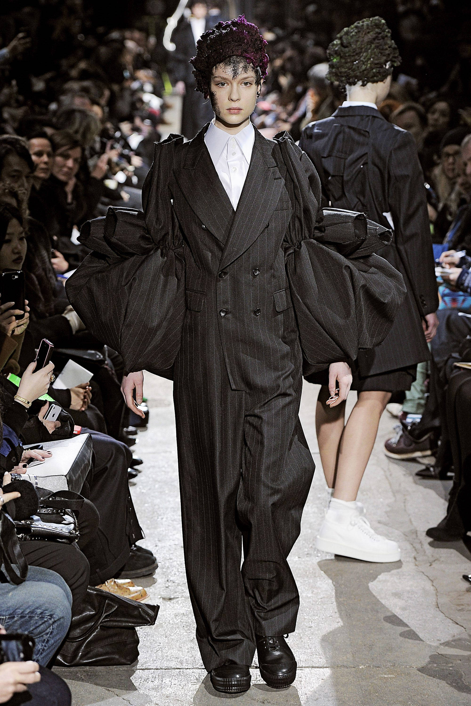 Comme Des Garçons 黑色蓬袖外套 壁紙 — 模特兒身穿誇張黑色細條紋西裝，搭配超大袖子和紫色花卉頭飾，在時裝展上走秀。