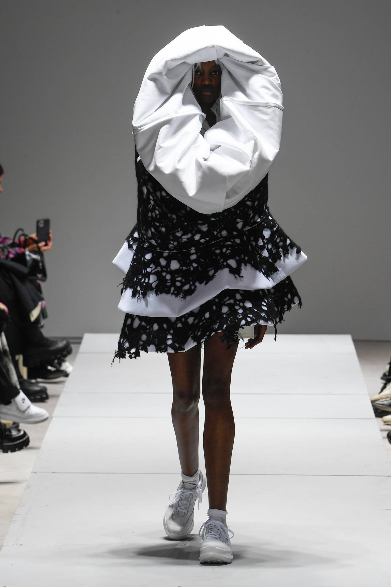 Comme des Garçons 前衛時裝頭飾 壁紙 — 一位模特兒穿著前衛黑白時裝，戴著大型垂墜兜帽和破損層疊裙，在走秀台上行走。