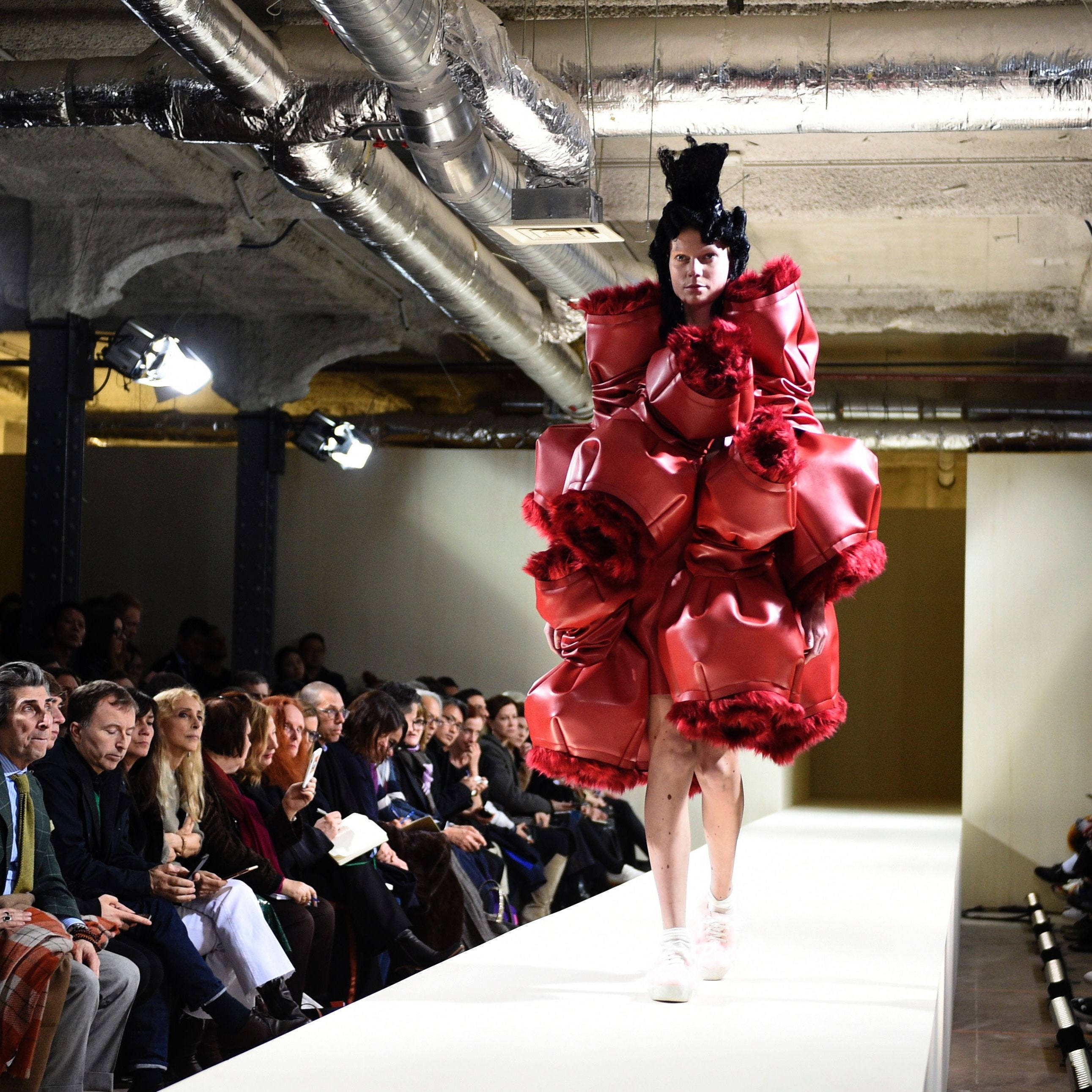 Comme Des Garçons紅色前衛皮革禮服 壁紙 — 模特兒穿著一件寬大的紅色毛邊連衣裙，在暴露的工業管道下走秀，觀眾在觀看。
