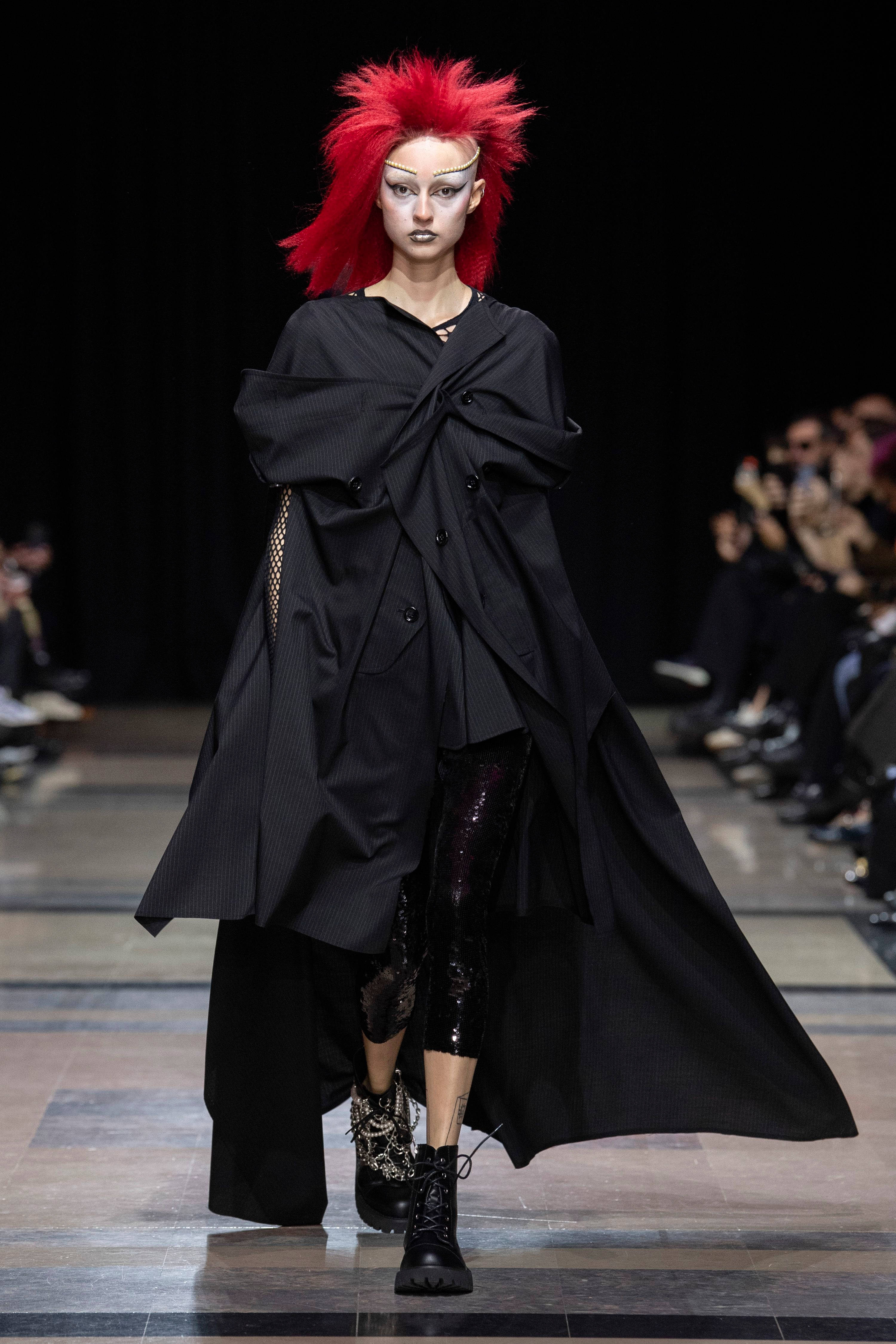 Comme Des Garçons 高級訂製黑色禮服 壁紙 — 一位擁有鮮紅色尖刺頭髮和大膽妝容的模特兒，自信地穿著一件戲劇性的超大黑色外套和閃亮緊身褲走在天橋上。