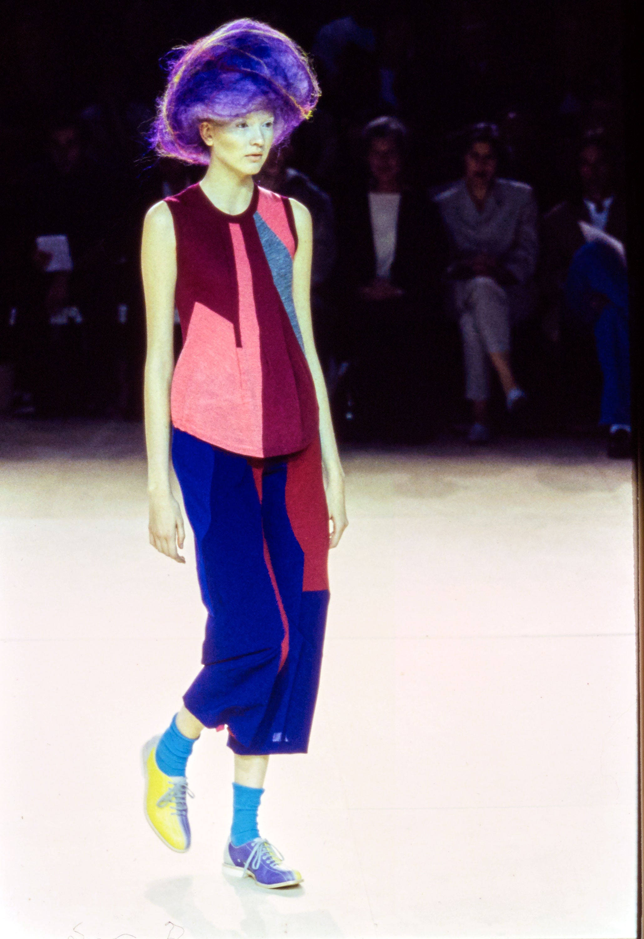 Comme Des Garçons 色彩幾何服裝 壁紙 — 一位擁有充滿活力的紫色頭髮的模特兒，在明亮的燈光下穿著大膽的幾何圖案服裝走秀。