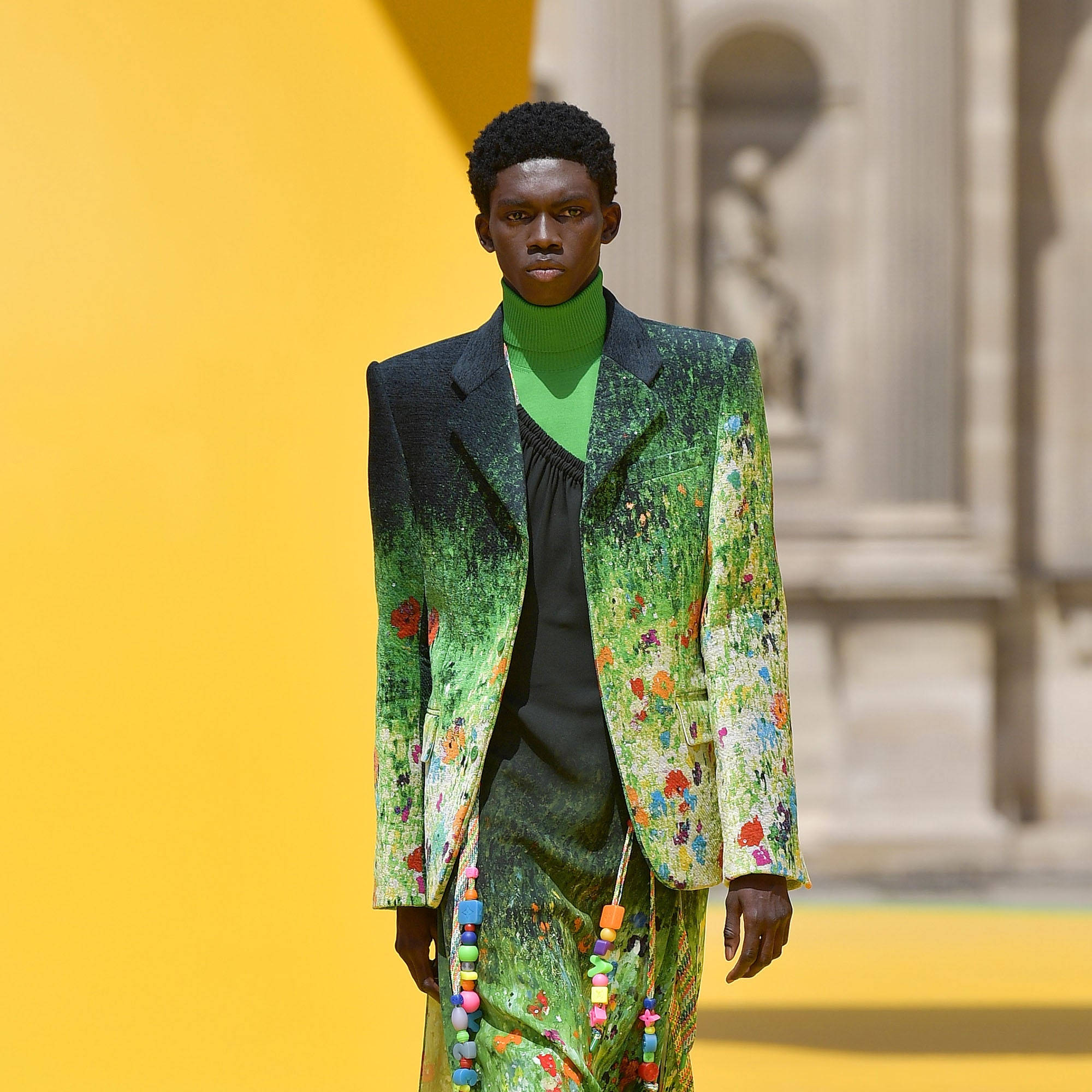Comme Des Garçons 綠色花卉西裝套裝 壁紙 — 一位模特兒自信地穿著充滿活力的綠色花卉圖案西裝，在帶有古典建築的鮮明黃色背景下走秀。