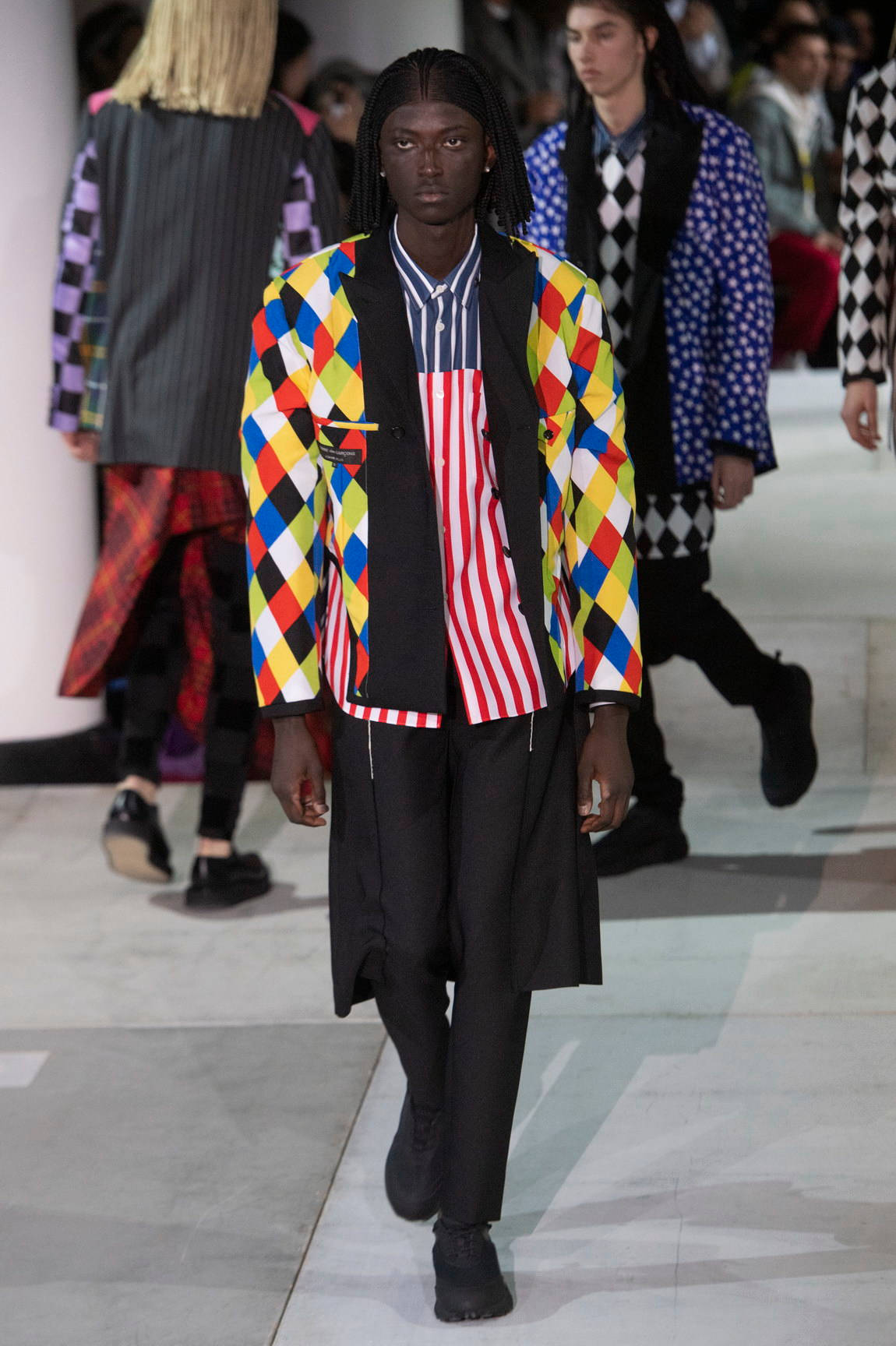 Comme Des Garçons彩色西裝外套 壁紙 — 一位模特兒在時裝天橋上走秀，身穿一件大膽的多色菱形圖案外套，內搭紅白條紋襯衫和黑色褲子。