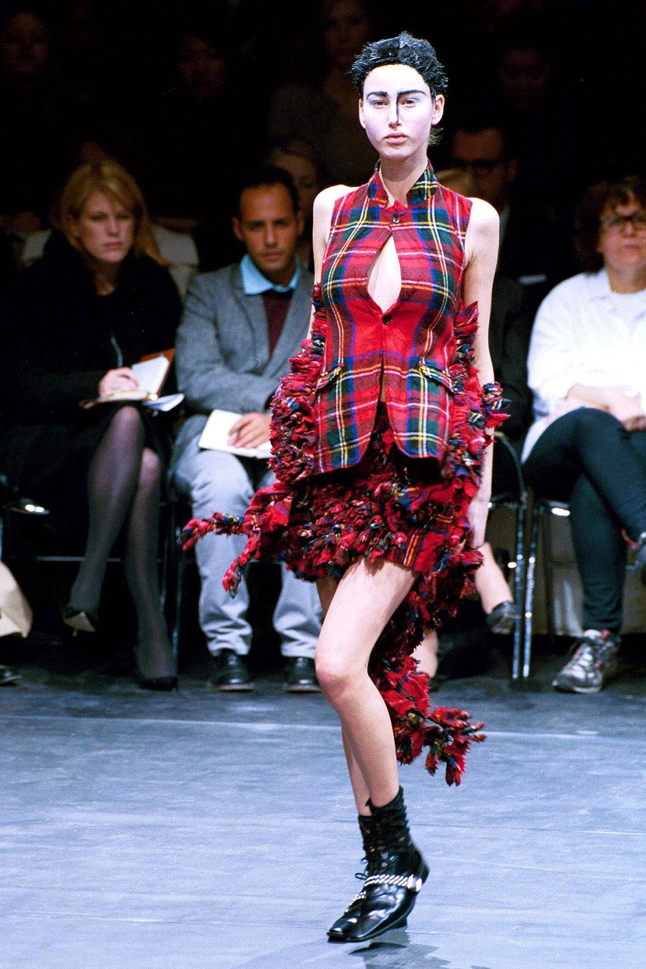 Comme Des Garçons紅色格紋短裙 壁紙 — 一位模特兒在明亮的舞台燈光下，穿著大膽的紅色格紋流蘇套裝和黑色靴子走在天橋上。