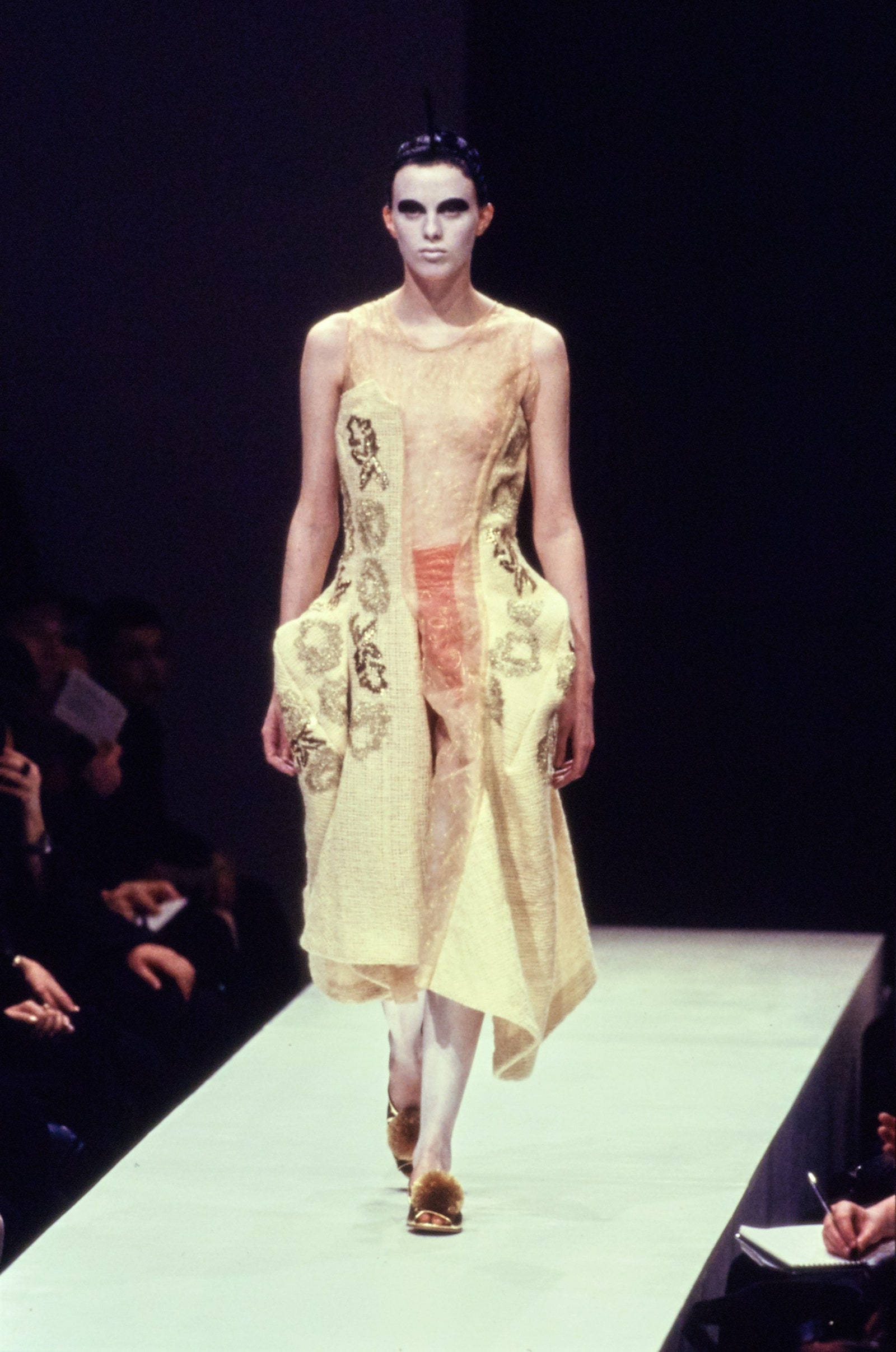 Comme Des Garçons 米色復古連衣裙 壁紙 — 一位妝容鮮明的模特兒穿著帶有花卉刺繡的質感淺黃色連衣裙和毛邊涼鞋走秀。