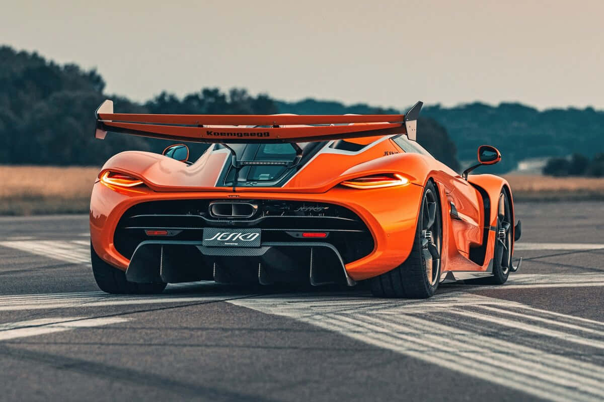  壁紙 — 從後方觀看，在日落時分的賽道上，一輛充滿活力的橙色Koenigsegg Jesko hypercar，背景是森林茂密的地平線。