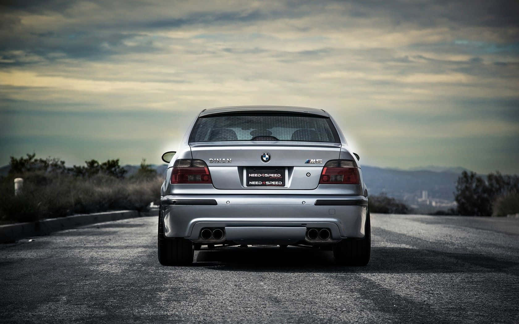 Bmw M5 桌布 壁紙 — 從後方觀看一輛銀色BMW M5房車在蜿蜒的道路上行駛，天空多雲，遠處有山丘。