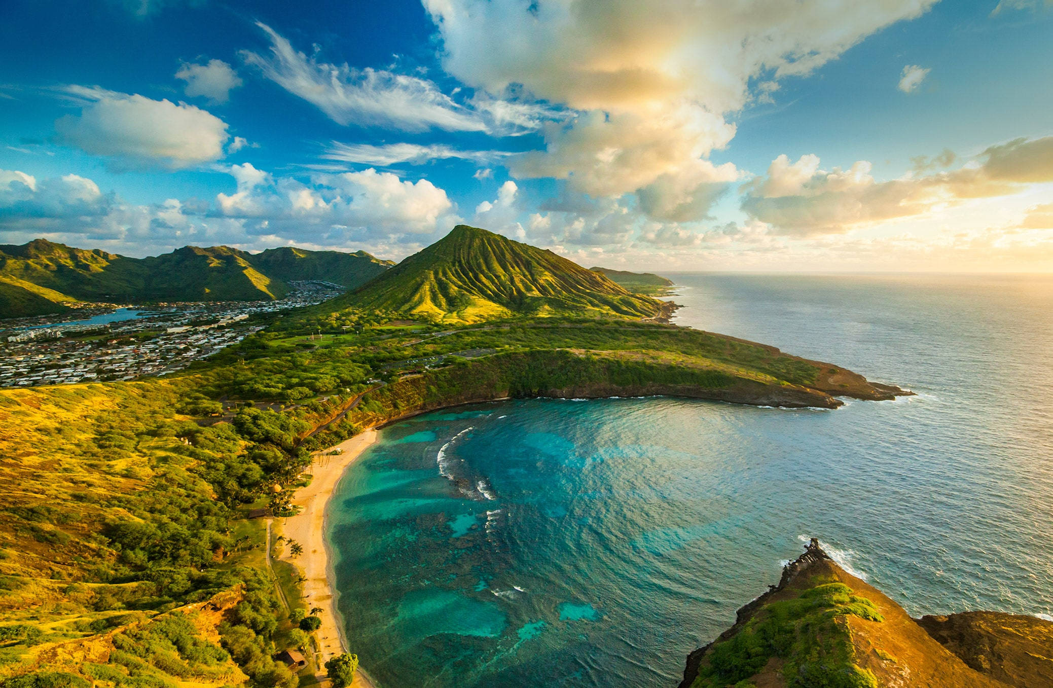 奧胡島壯麗日落風景 壁紙 — 從空中俯瞰一個鬱鬱蔥蔥的綠色火山錐，俯瞰著一個碧綠的海灣，有沙灘和沿海小鎮，天空湛藍，雲朵點綴。