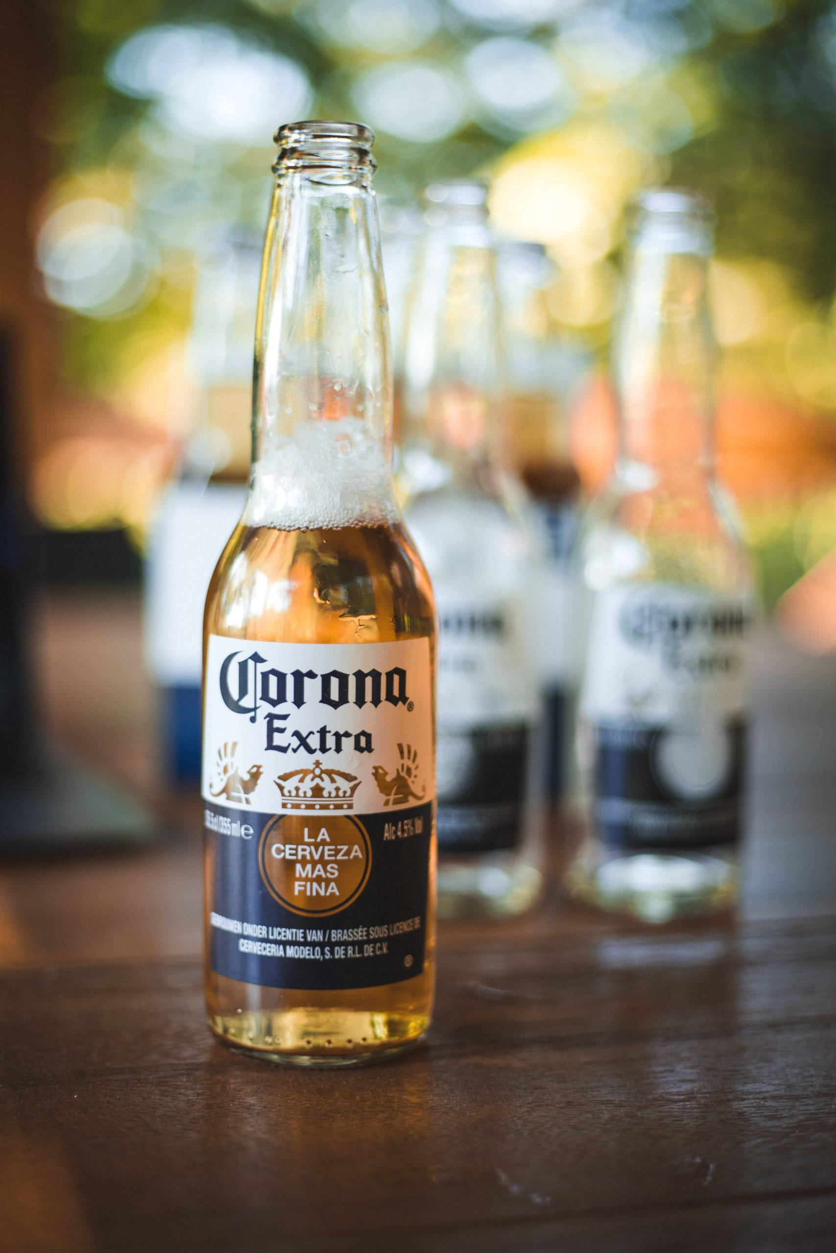 Corona Extra墨西哥啤酒廣告 壁紙 — 一瓶冰凍的Corona Extra啤酒瓶，帶有水珠，放在木桌上，背景有模糊的瓶子。