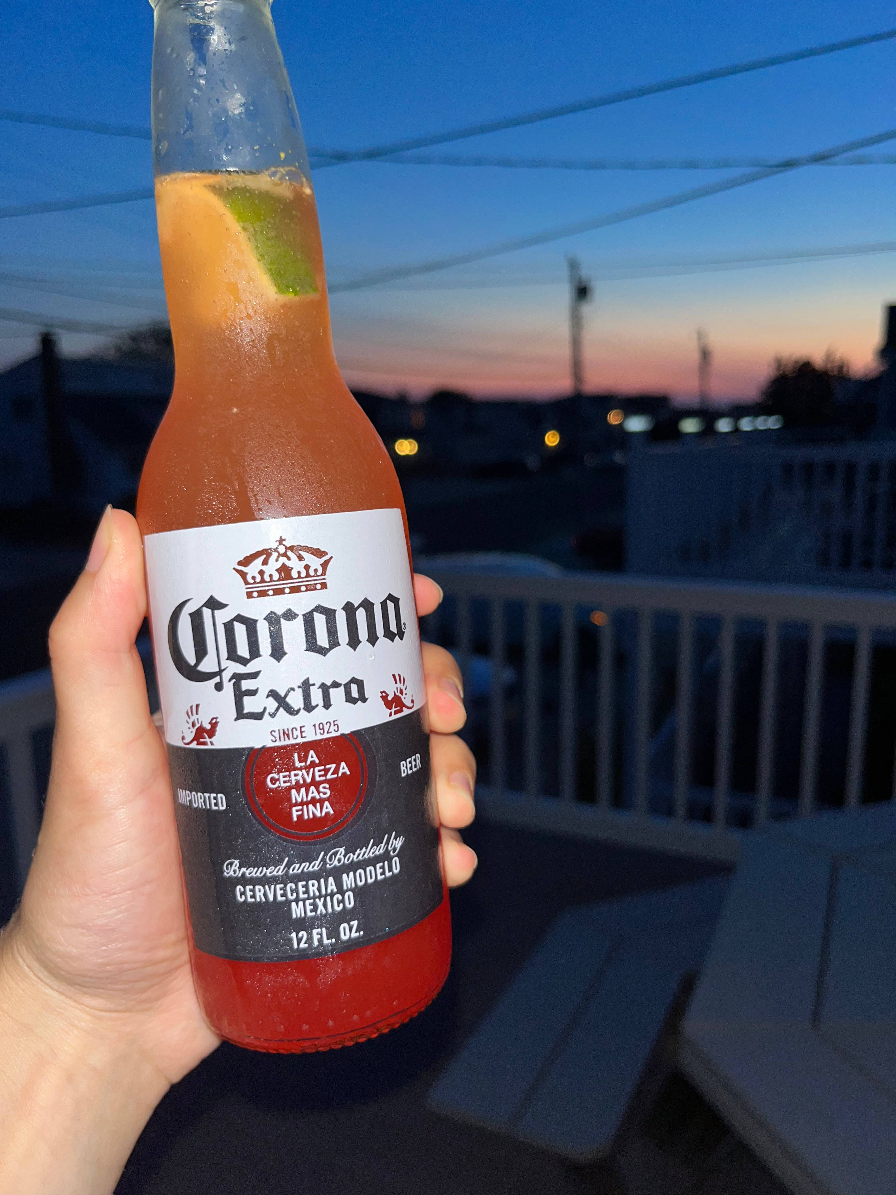 Corona Extra日落時分 壁紙 — 日落時分，一隻手在可俯瞰城市景觀的陽台上拿著一杯加了青檸的冰凍Corona Extra啤酒。