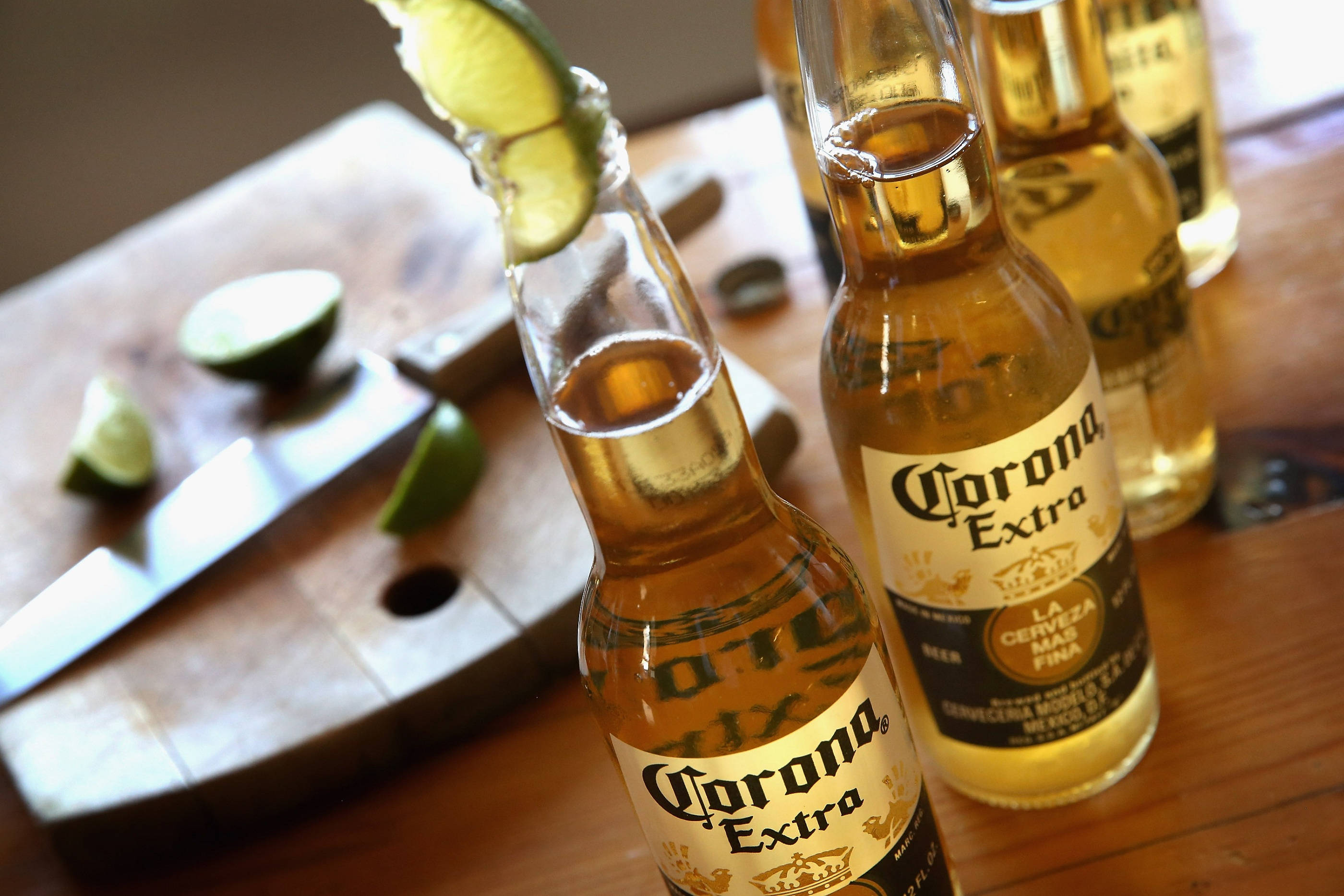 Corona Extra綠檸檬暢飲風景 壁紙 — 金色 Corona Extra 啤酒瓶配青檸角放在木桌上，旁邊有砧板放著青檸片和一把刀。
