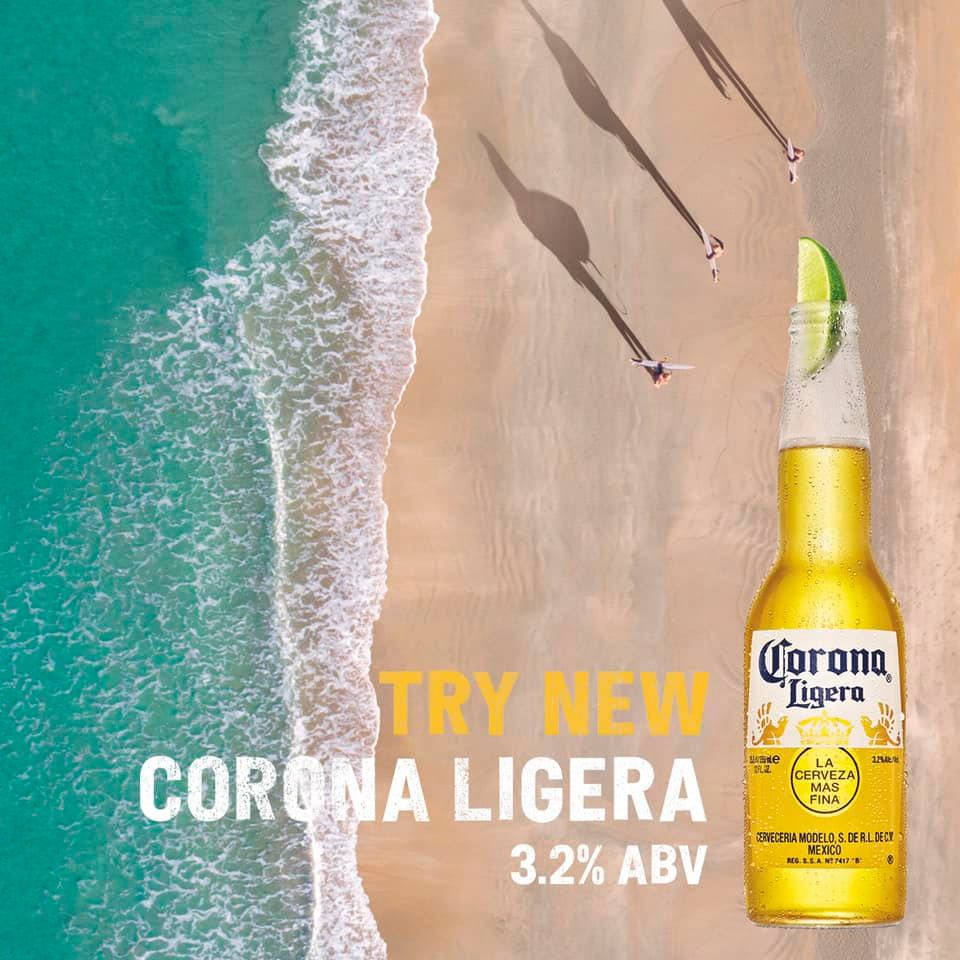 Corona Ligera新啤酒海報 壁紙 — 一瓶Corona Ligera啤酒連青檸角放在沙灘上，旁邊有碧綠海浪和玩長板的人的剪影。