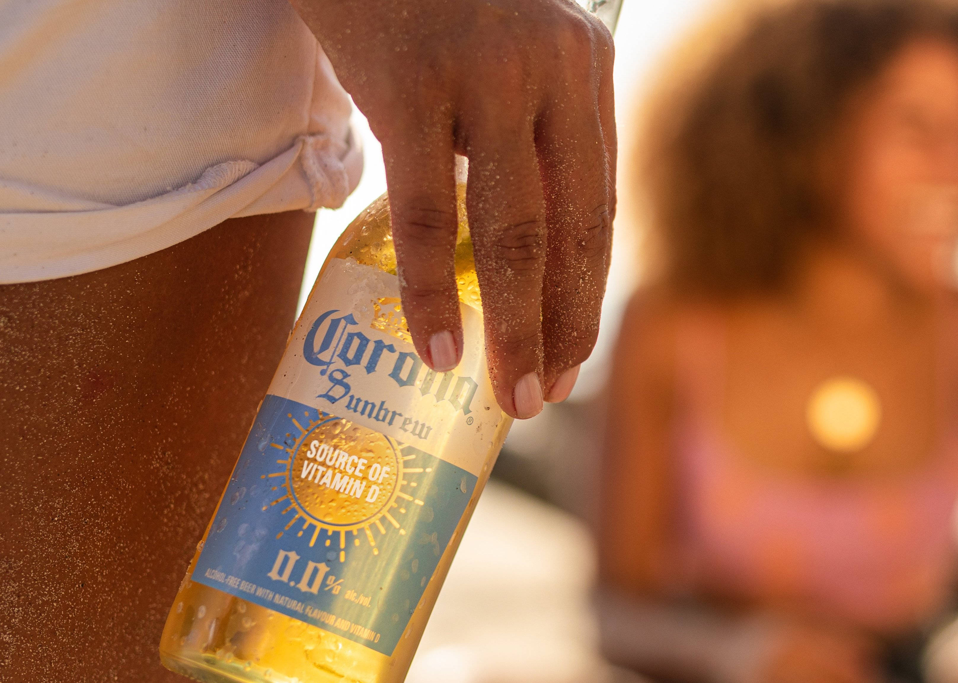 Corona Sun Brew 海灘版 壁紙 — 一隻帶沙的手拿著Corona Sunbrew瓶子，背景是陽光明媚的海灘，背景中有一個模糊的人影。