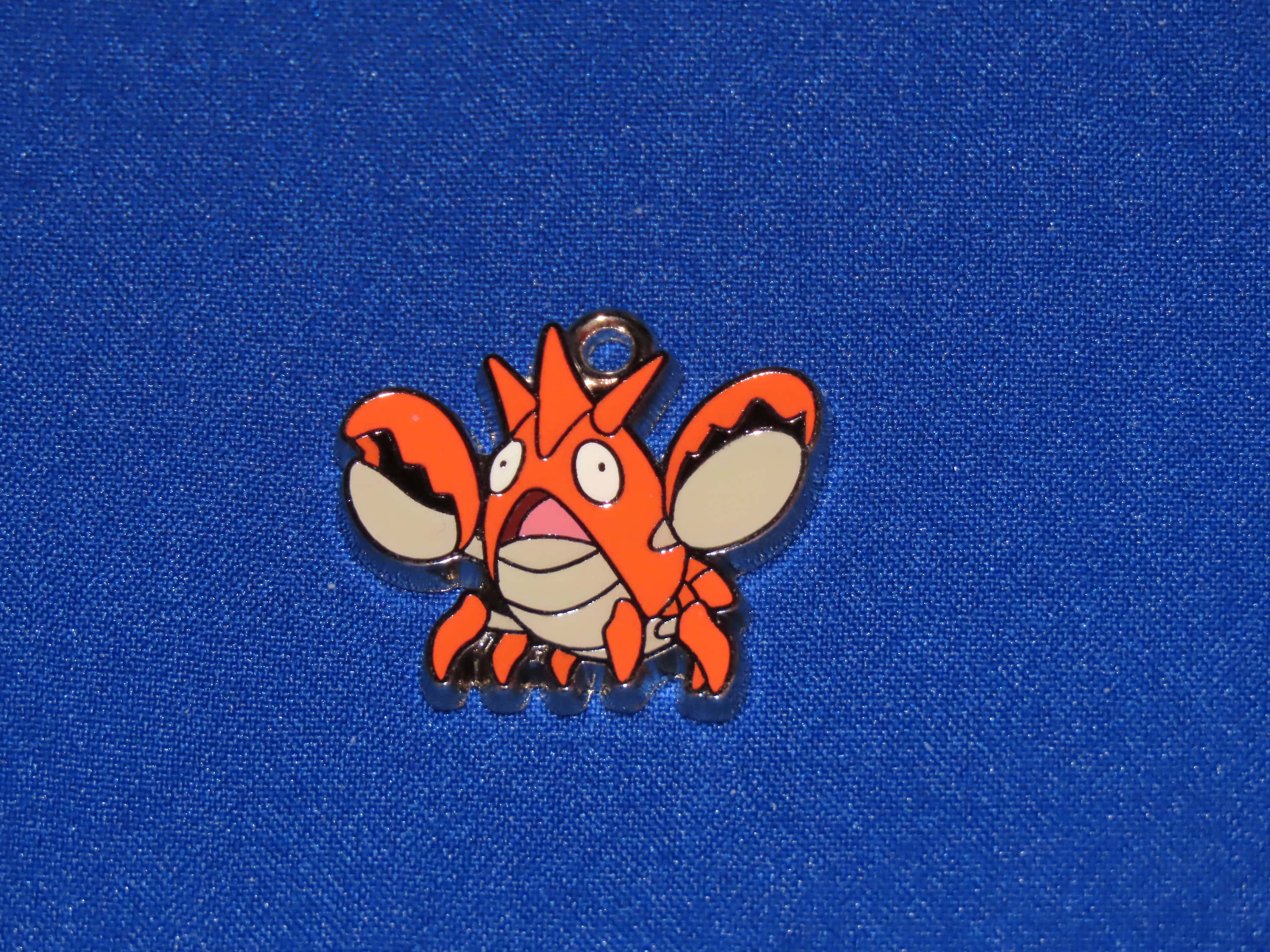 Corphish吊飾 壁紙 — 一枚亮橙色嘅Krabby Pokémon琺瑯別針，有白色眼睛同米色爪，放喺有紋理嘅皇家藍色布料背景上。