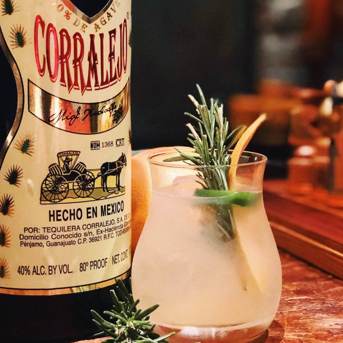 墨西哥雞尾酒 Corralejo 龍舌蘭酒烈酒 壁紙 — 一杯冰涼的龍舌蘭雞尾酒，飾以迷迭香和柑橘，放在溫暖的木吧檯上，旁邊是一瓶 Corralejo。