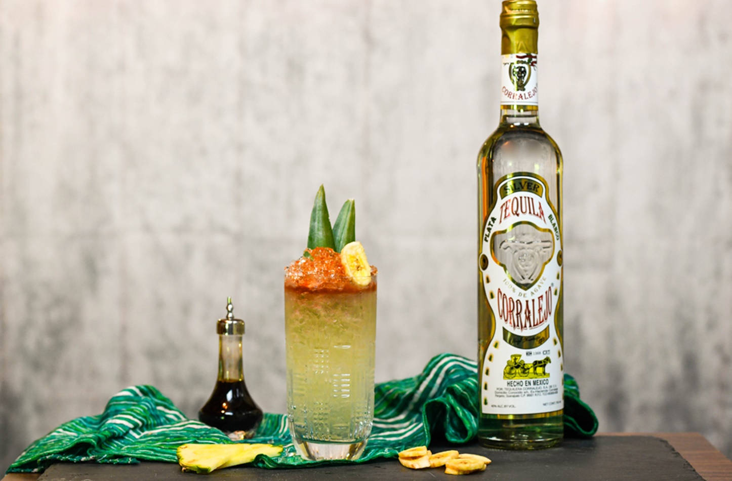 獨特 Corralejo 龍舌蘭酒提基雞尾酒 壁紙 — 一杯充滿活力的龍舌蘭雞尾酒，以香蕉和龍舌蘭裝飾，旁邊放著一瓶Corralejo龍舌蘭酒，置於綠色布料上，背景為紋理牆壁。