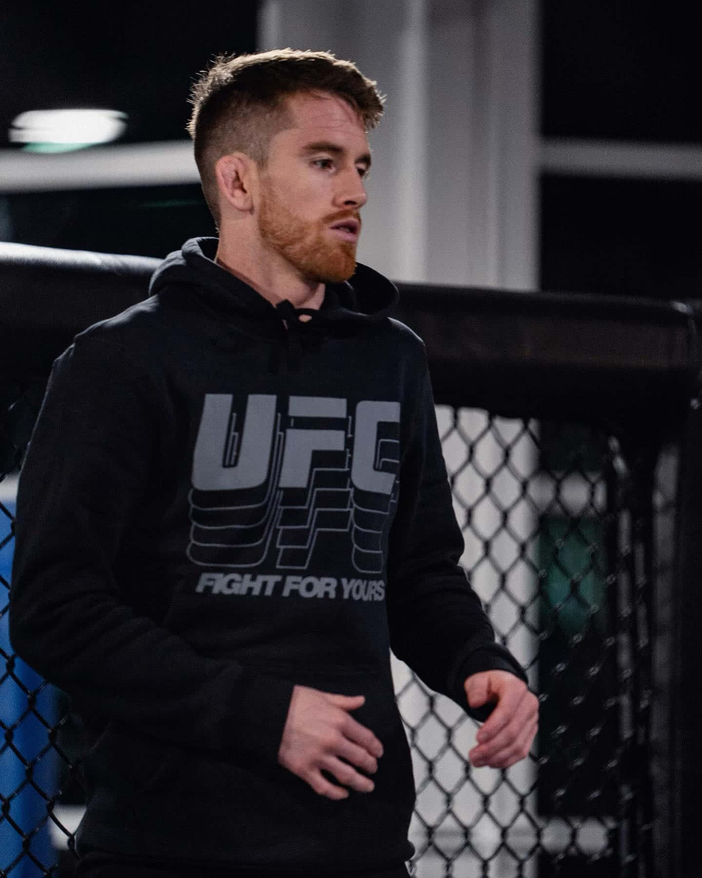 Cory Sandhagen穿UFC連帽衫 壁紙 — 一位專注的男性拳手穿著黑色UFC連帽衫，夜晚站在昏暗的八角籠內。