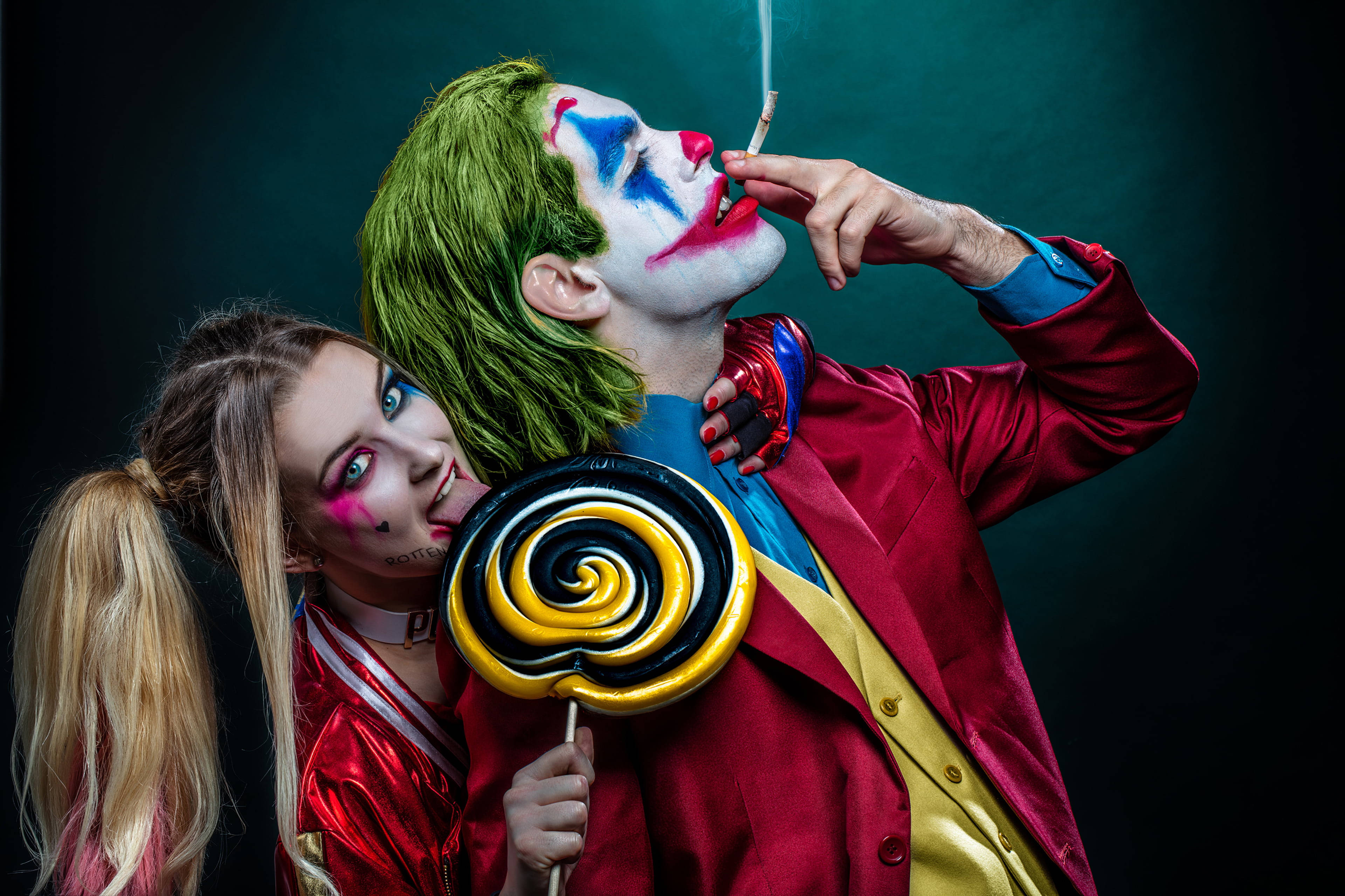 小丑與哈莉·奎茵 cosplay 壁紙 — 一個綠髮小丑妝的男人抽煙，而一個紮辮子的女人在深藍綠色背景下拿著一個巨大的漩渦棒棒糖。
