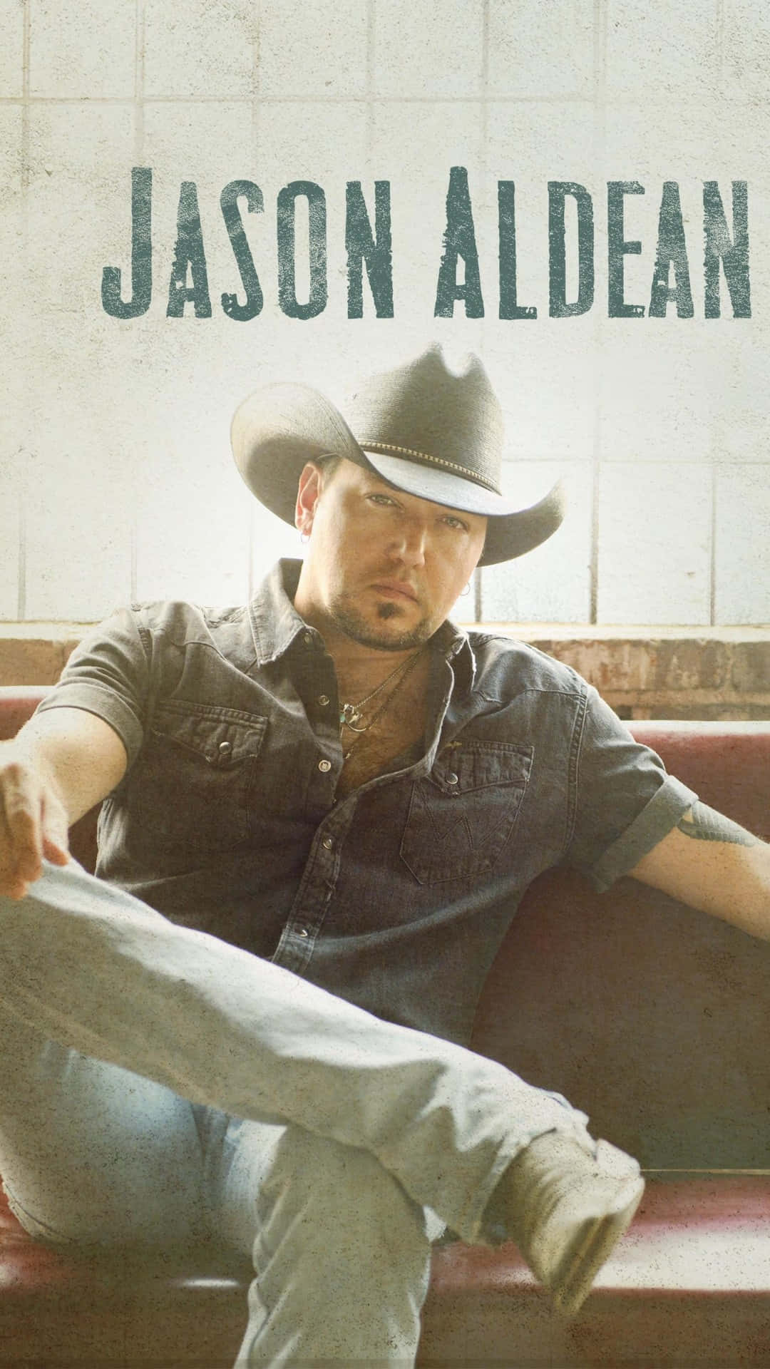  壁紙 — 鄉村歌手 Jason Aldean 戴著牛仔帽、穿著牛仔襯衫和牛仔褲，休閒地坐在紅色長椅上，背景是瓷磚牆。