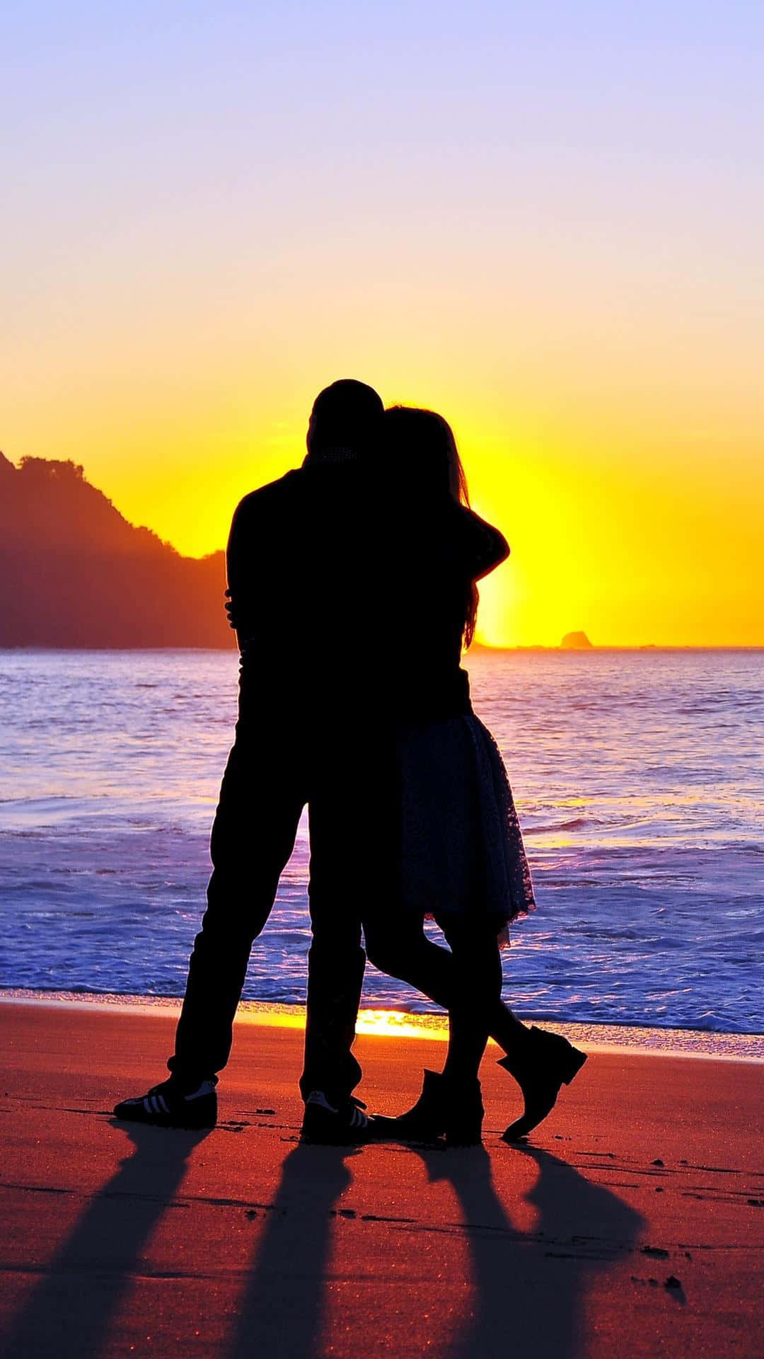  壁紙 — 一對情侶在海灘上以剪影相擁，夕陽在他們身後落下，投下長長的影子。