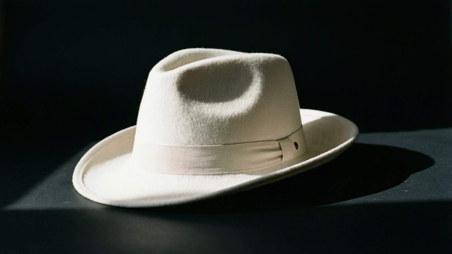 White Hat Minimalist 壁紙 — 一頂奶油色軟呢帽搭配相配的帶子，在黑暗背景上的戲劇性聚光燈下靜置。