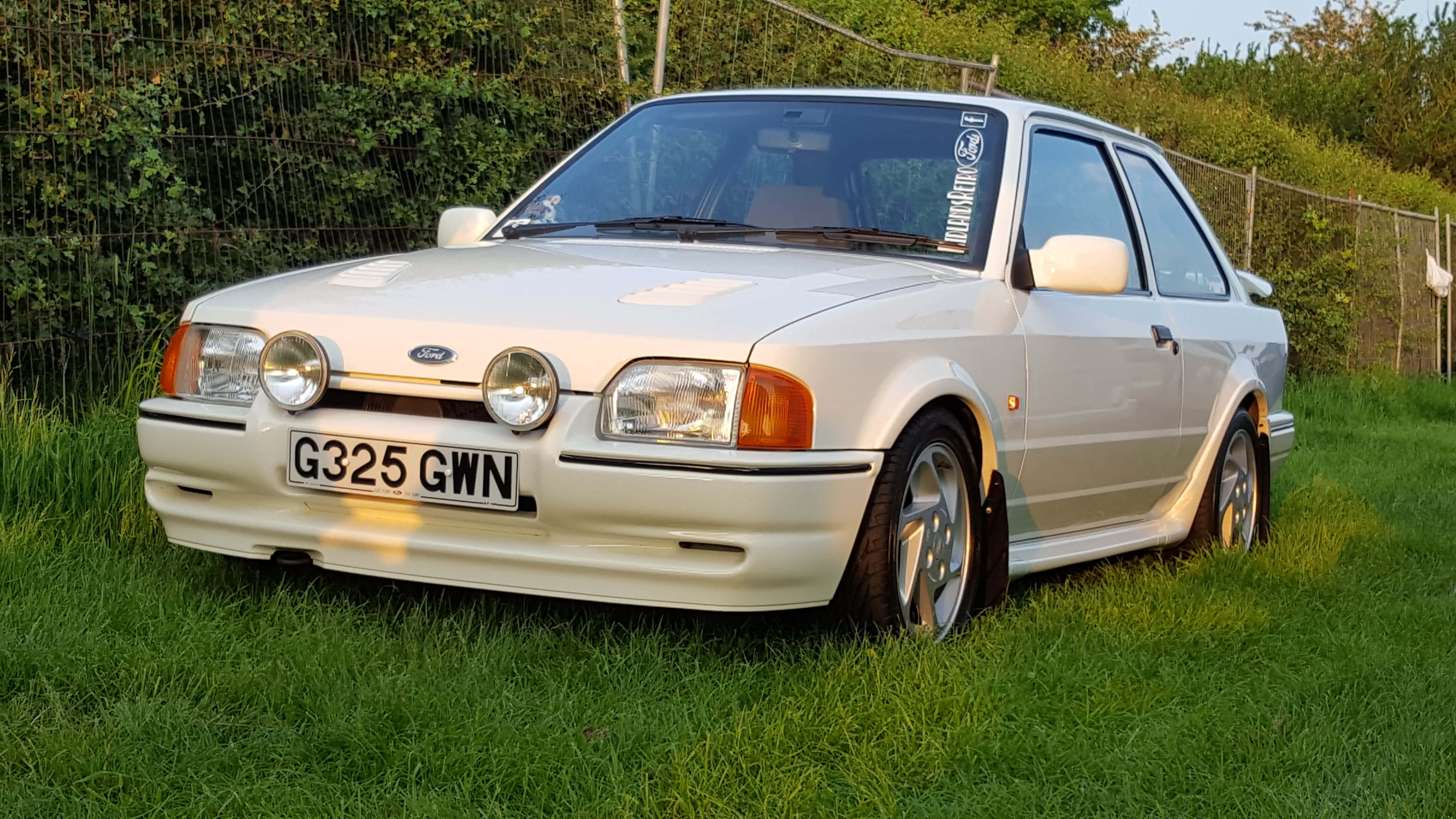 Ford Escort 桌布 壁紙 — 一架奶油色福特 Sierra Cosworth 停泊在草地上，後面有灌木叢和籬笆，在溫暖的傍晚光線下。