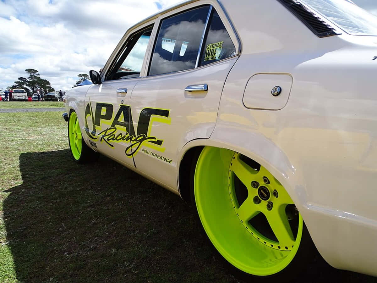 Mazda Rx-3 桌布 壁紙 — 一輛奶油色改裝轎車，配備螢光綠色車輪和PAC Racing貼紙，停在多雲天空下的草地上。