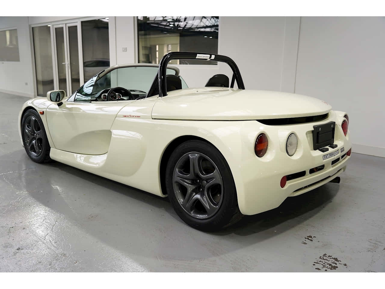 Renault Sport Spider 桌布 壁紙 — 一輛完美的奶油色跑車，帶有黑色飾邊和防滾架，停放在室內的灰色地板上。