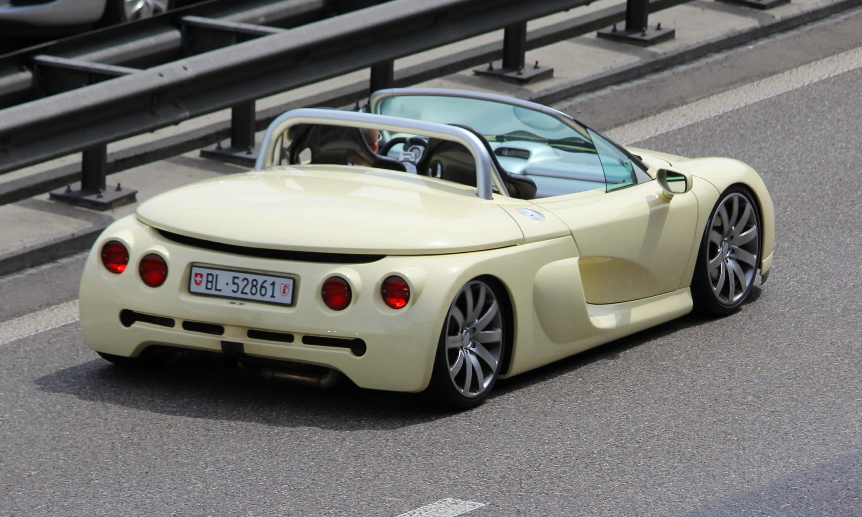 Renault Sport Spider 桌布 壁紙 — 一輛奶油黃色開篷跑車，帶有紅色尾燈，在陽光普照下，沿著有護欄的高速公路行駛。