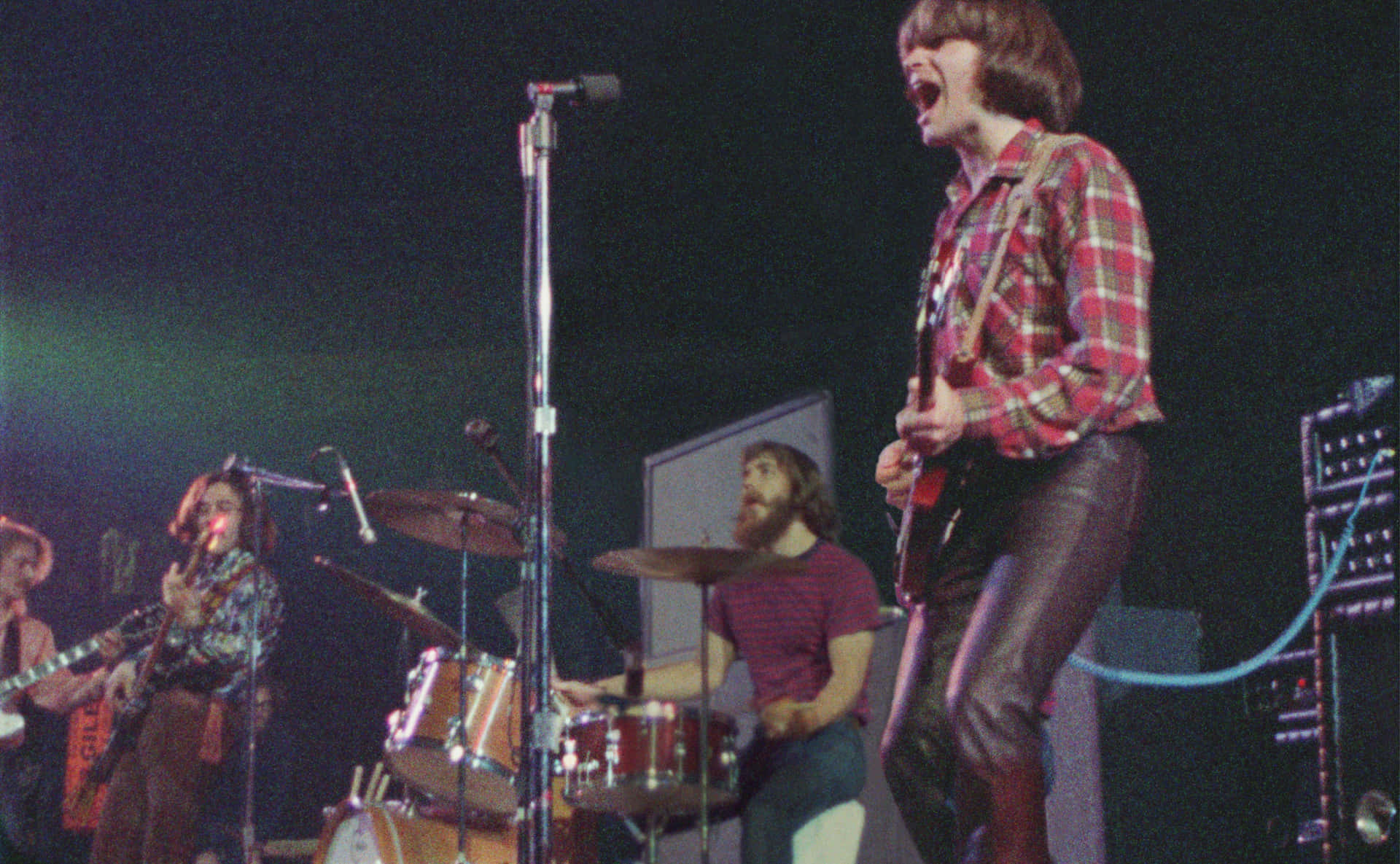 Creedence Clearwater Revival 演出現場 壁紙 — 一支復古搖滾樂隊在昏暗燈光下於舞台上充滿活力地表演，一位穿著格仔恤衫的結他手正熱情地唱歌。