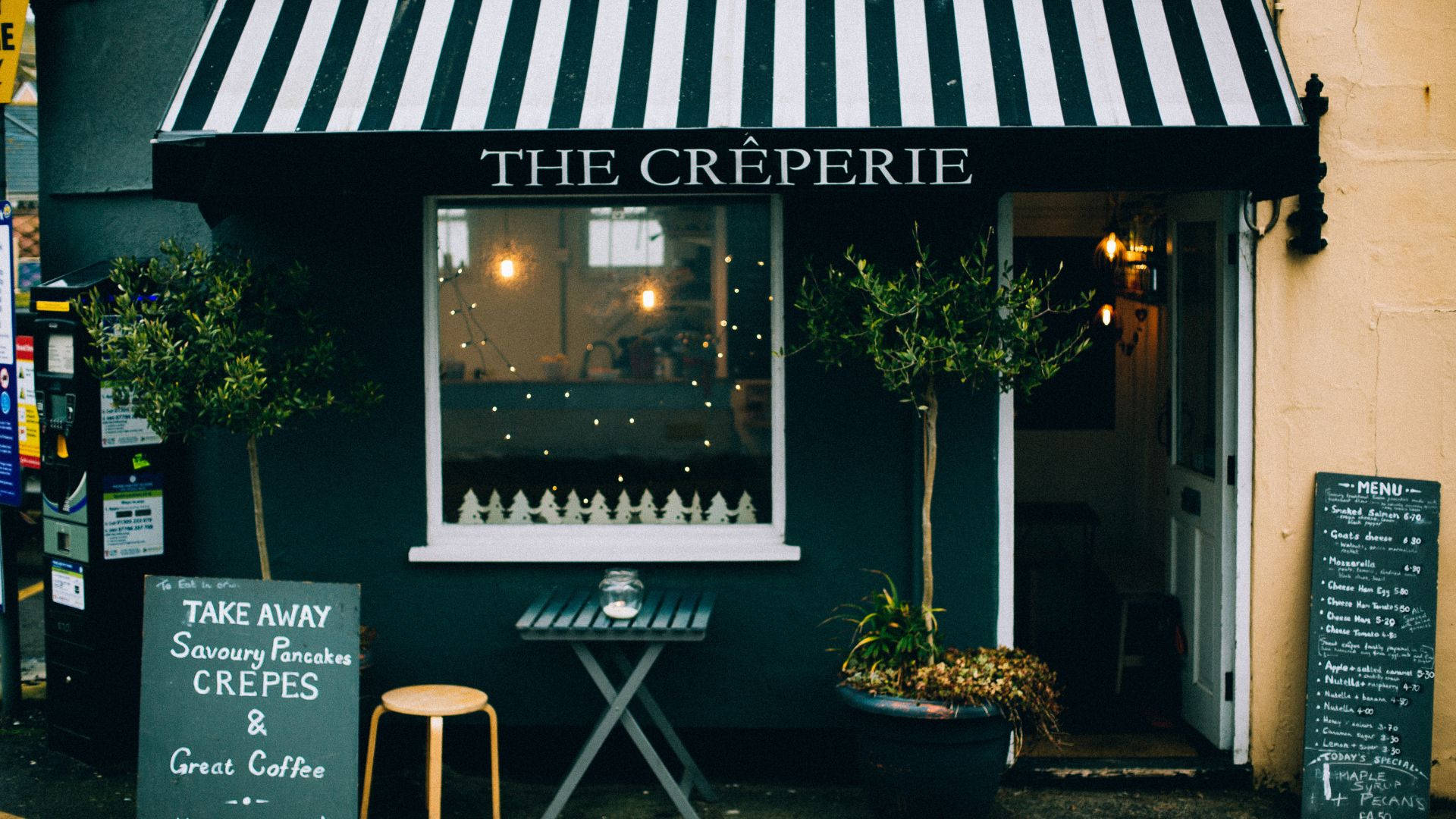Creperie 店面黑白條紋遮陽篷 壁紙 — 一間舒適的可麗餅店，設有黑白條紋遮陽篷，店內散發溫暖燈光，入口兩旁擺放盆栽植物。
