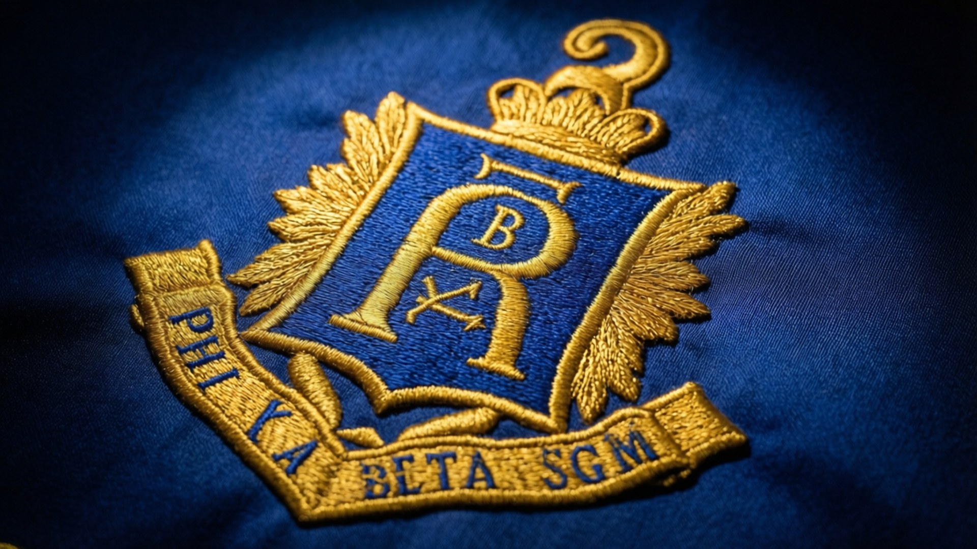 藍色布料上金色刺繡的 Phi Kappa Beta SGM 徽章，帶有月桂葉翅膀和皇冠