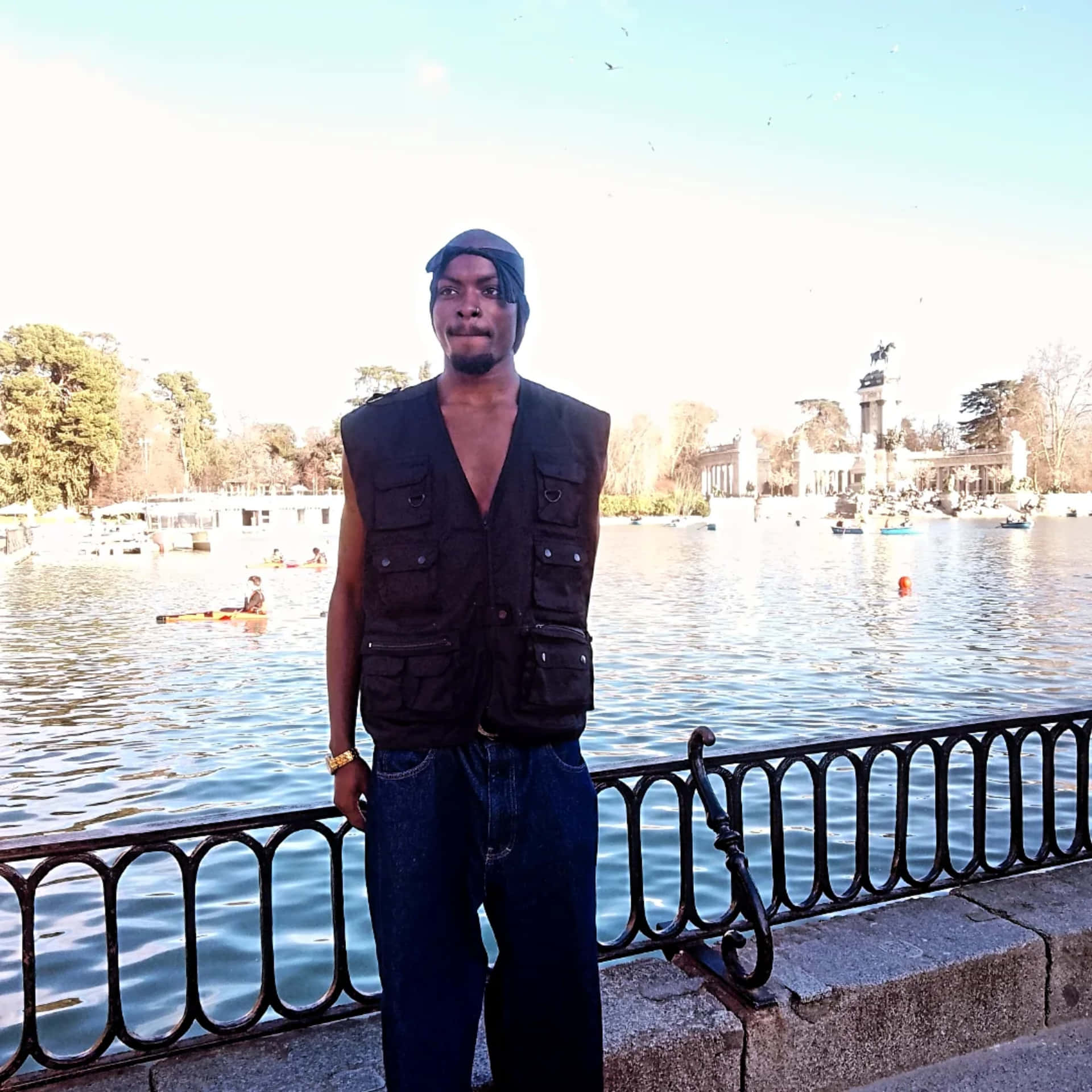 身穿黑色背心和牛仔褲的男子站在湖邊，背景有划獨木舟的人和紀念碑