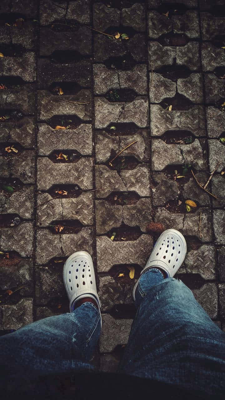 穿著Crocs，邁入舒適與時尚 壁紙 — 第一人稱視角，雙腳穿著白色Crocs和藍色牛仔褲，站在佈滿落葉的風化石鋪路面上。