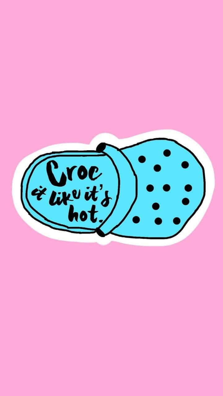 體驗Crocs帶來的舒適與時尚 壁紙 — 一個卡通風格的藍色 Croc 鞋貼紙，上面寫著「Croc it like it's hot」，背景為粉紅色。