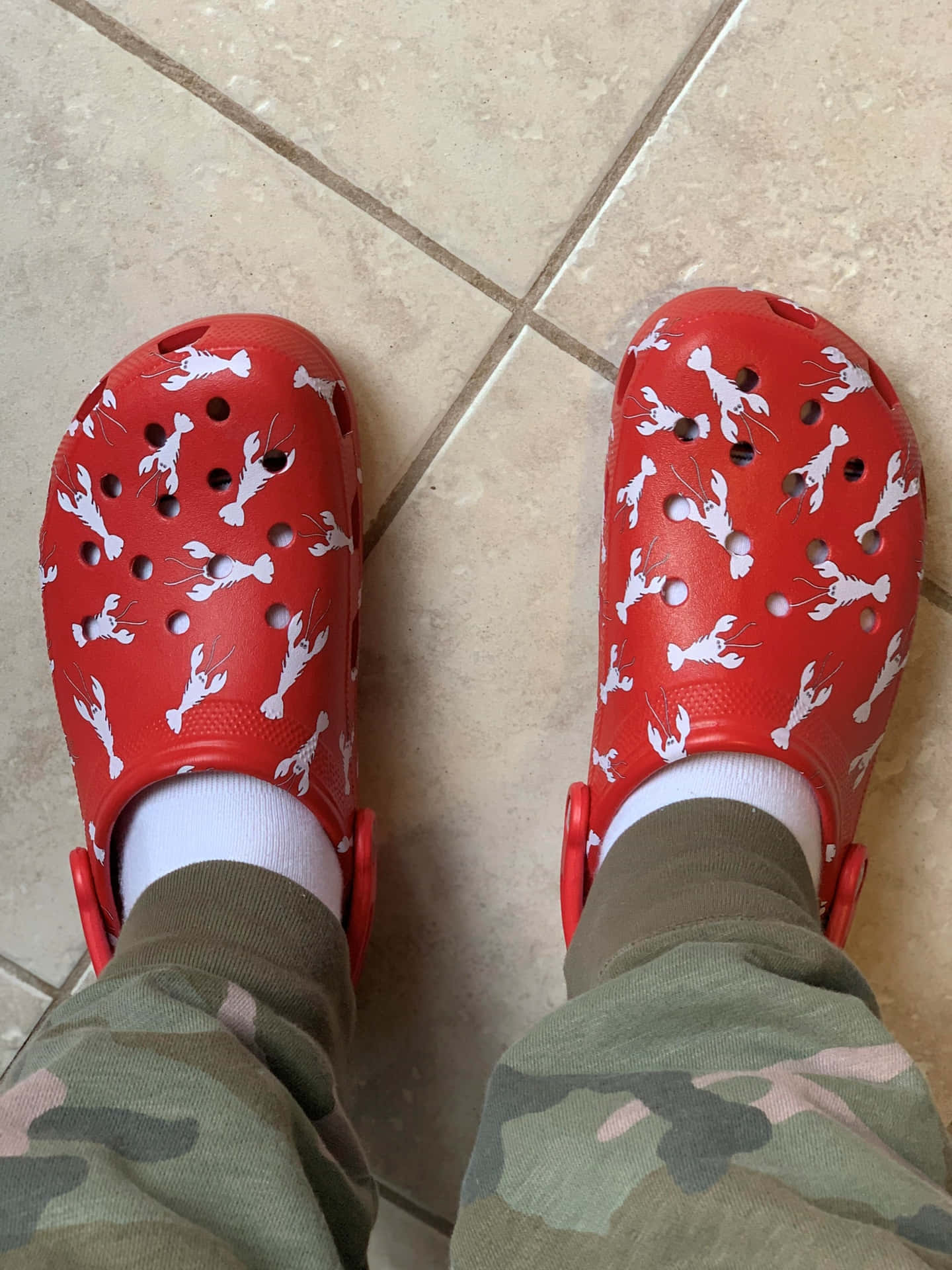 米色瓷磚地板上的紅色Crocs鞋，帶有白色龍蝦圖案，搭配迷彩褲和白色襪子。