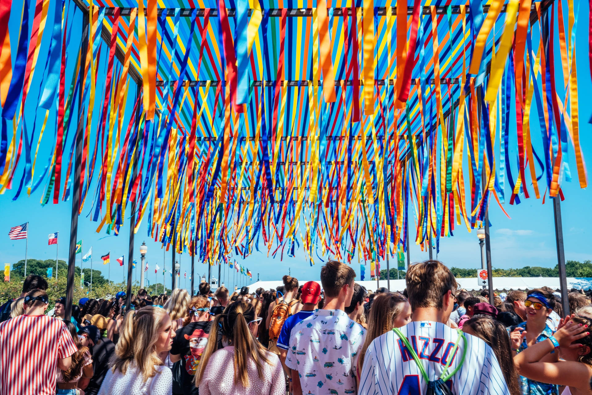 叫上朋友，Lollapalooza來了！ 壁紙 — 人群在色彩繽紛的彩帶天篷下行走，背景是清澈的藍天。