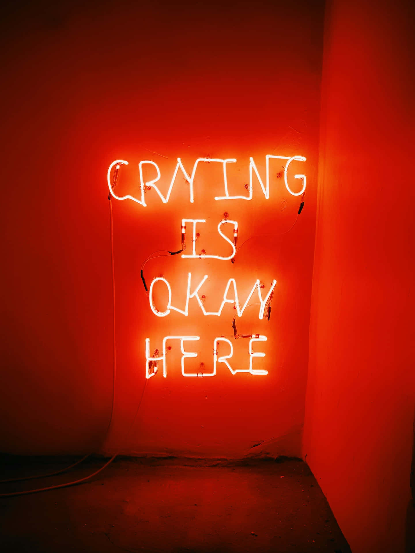  壁紙 — 一個發光的白色霓虹燈牌，上面寫著 crying is okay here，在昏暗的房間中，將溫暖的紅光投射在樸素的牆壁上。