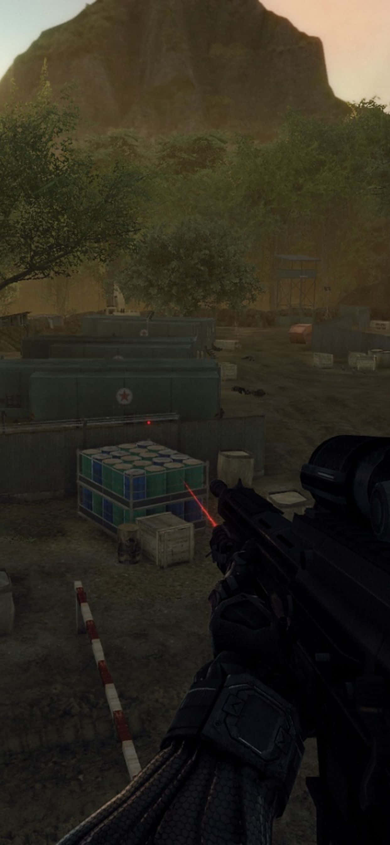 Crysis Warhead 遊戲在 iPhone Xs Max 上呈現的震撼視覺 壁紙 — 第一人稱視角，一名士兵在黃昏時分用紅色激光瞄準器瞄準一個被樹木和岩石山包圍的軍事基地。