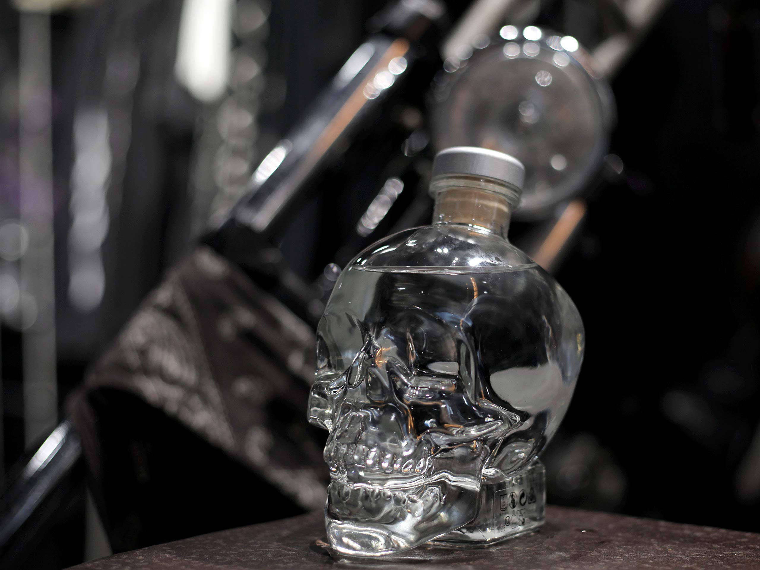 Crystal Head伏特加於工業環境中 壁紙 — 一個透明骷髏形狀的伏特加酒瓶放在黑暗的表面上，背景有模糊的電單車零件。