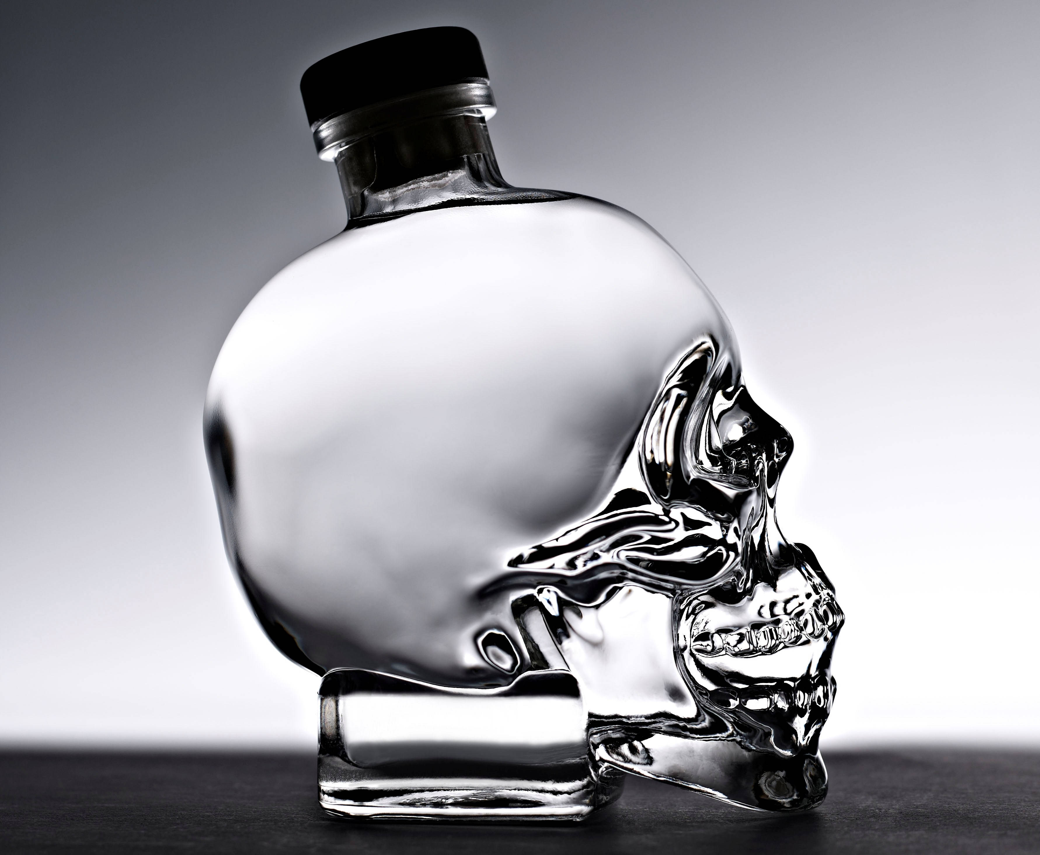 Crystal Head伏特加瓶的精緻工藝 壁紙 — 一個透明玻璃骷髏形狀的瓶子，帶有黑色蓋子，放在深色表面上，背景為漸變灰色。