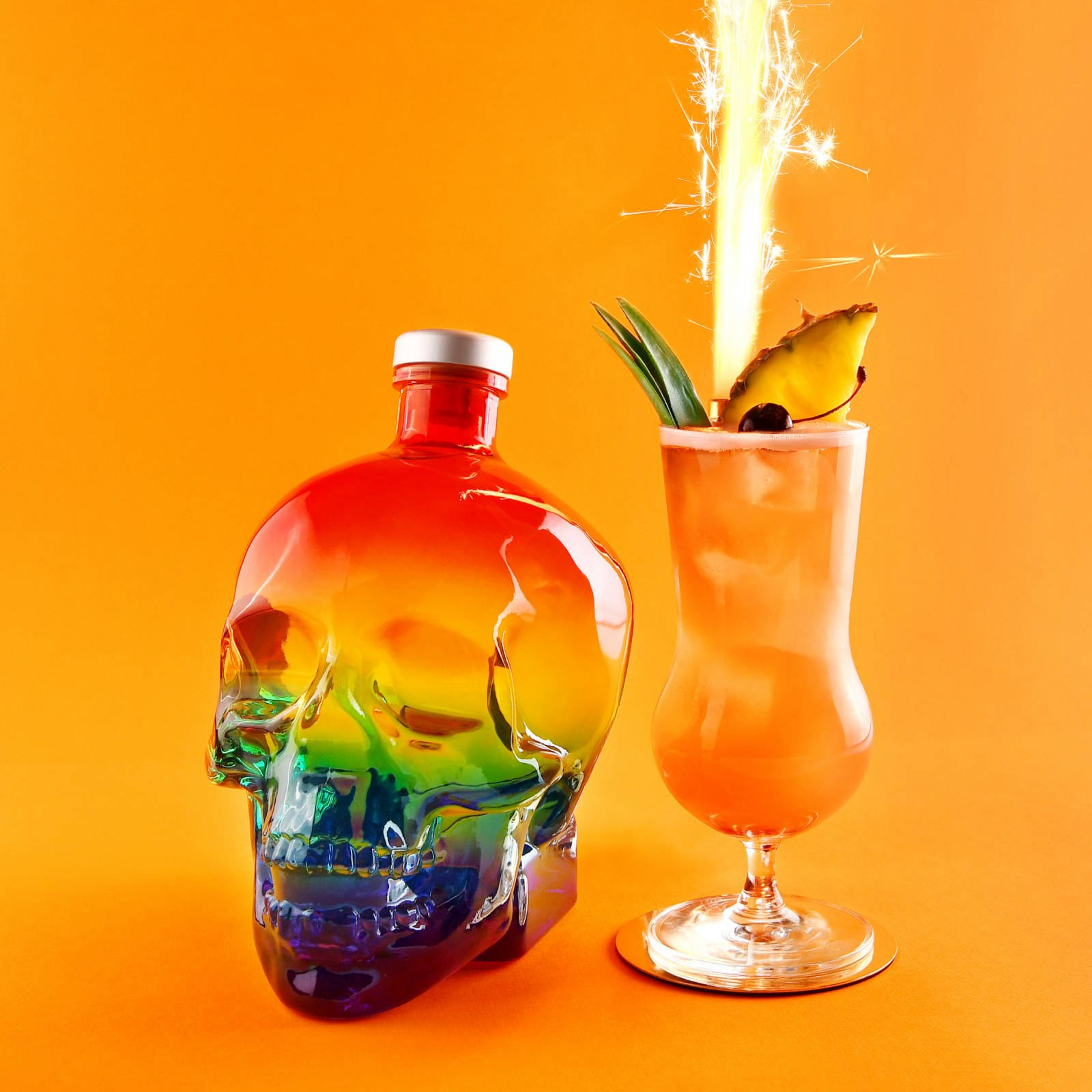 以Crystal Head伏特加調製的熱帶飲品 壁紙 — 彩虹骷髏樽同熱帶雞尾酒，配上煙花裝飾，襯托鮮豔橙色背景。