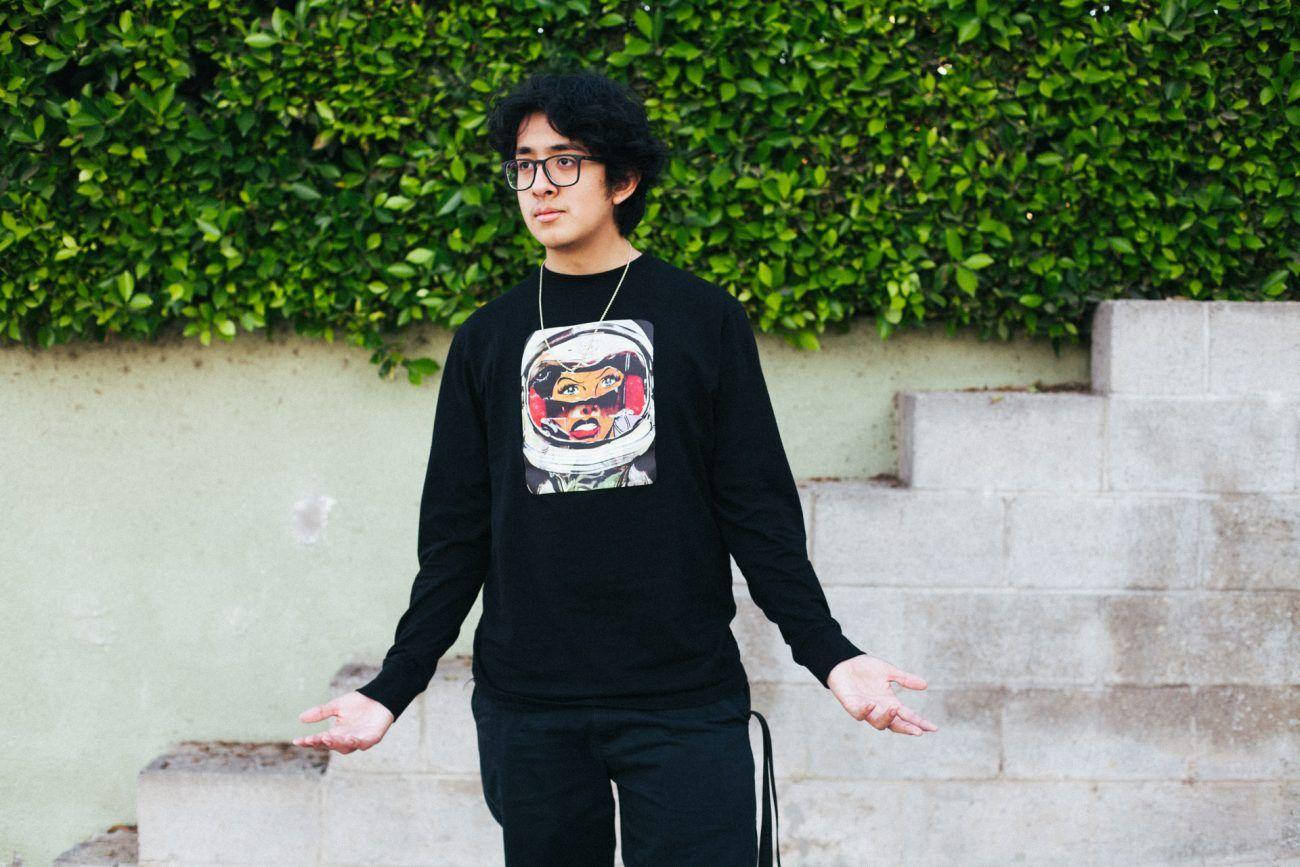 戴眼鏡、身穿太空人運動衫的年輕男子站在戶外，靠近混凝土台階和綠色籬笆。
