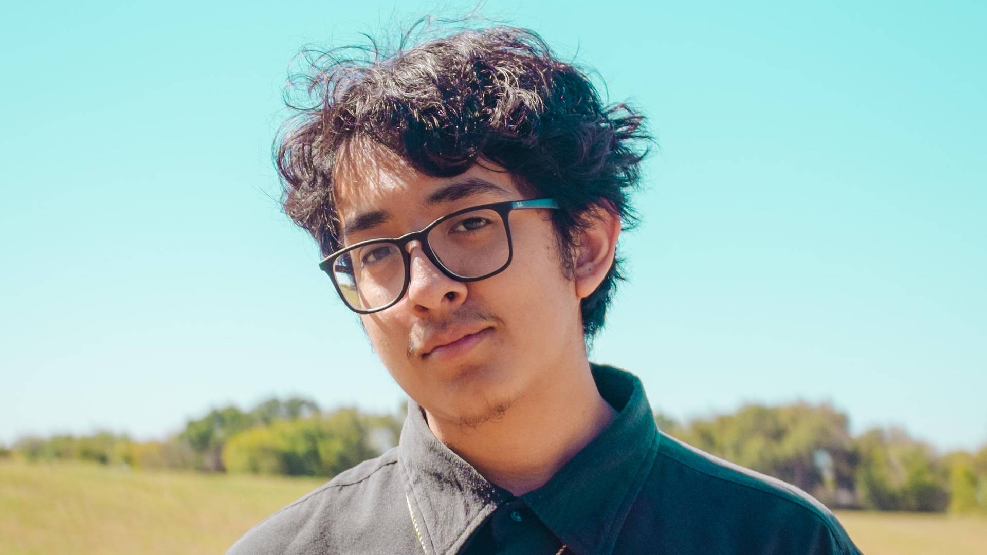 戴眼鏡的年輕男子站在陽光明媚的田野中，背景有樹木。