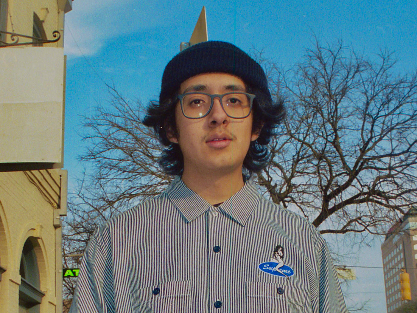 戴眼鏡和冷帽的年輕男子站在城市街道上，周圍有光禿的樹木和建築物。