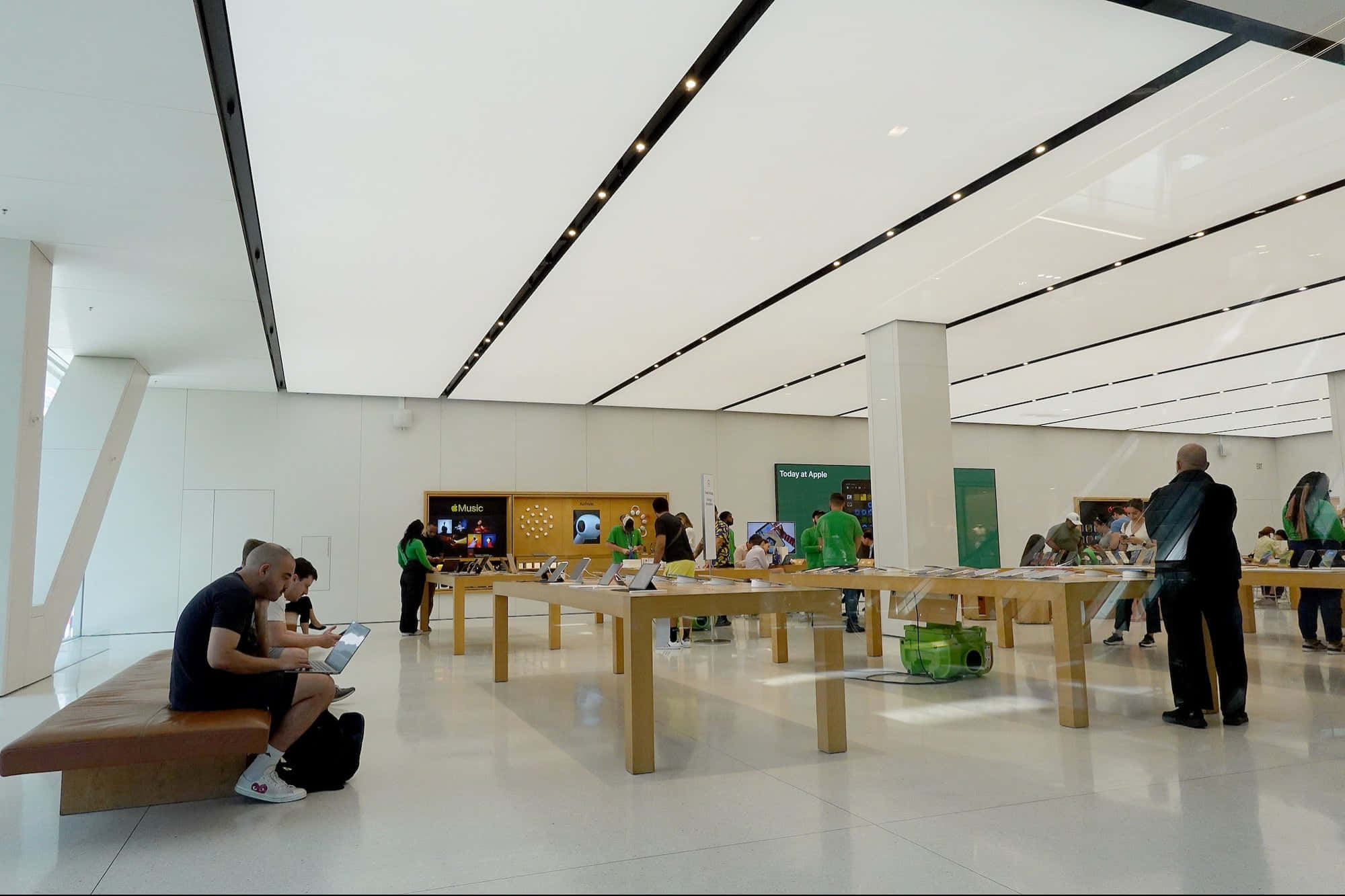 Apple Store 桌布 壁紙 — 顧客瀏覽時尚木桌上展示的Apple產品，店舖明亮簡約，高樓底配現代燈光。